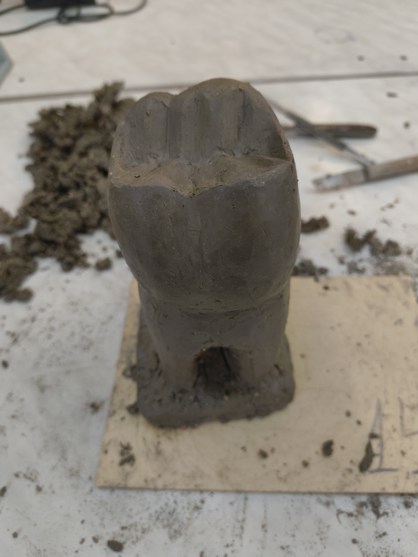 A close-up shot shows a clay model of a molar tooth, positioned on a square piece of cardboard. Clumps of clay are scattered to the left of the model. Two metal sculpting tools are visible in the background to the right. The scene appears to be a workspace with clay residue on the surrounding surface. There is no evidence of the location being Sofia, Bulgaria. There is no evidence of people.