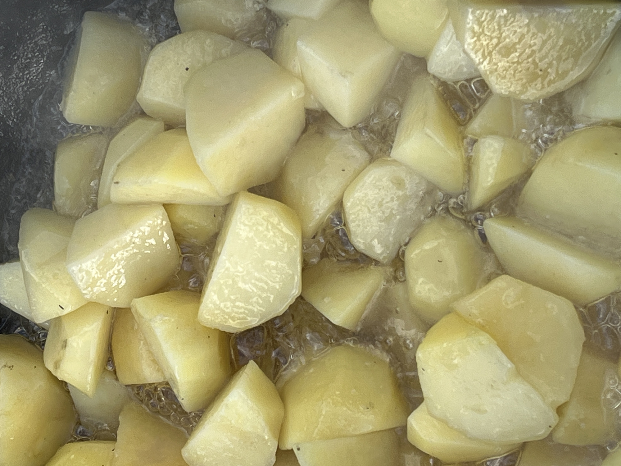 The image shows a close-up, overhead view of numerous potato chunks submerged in bubbling water within a pot. The potatoes are cut into irregular, bite-sized pieces and appear to be in the process of boiling. Some pieces are slightly translucent, indicating they are softening. The water is actively boiling, with visible bubbles rising around the potatoes. The lighting is bright, highlighting the smooth, pale yellow surface of the potato pieces and the shimmering water. There is no discernible indication of the location within Il'ino, Russia, or any people present.