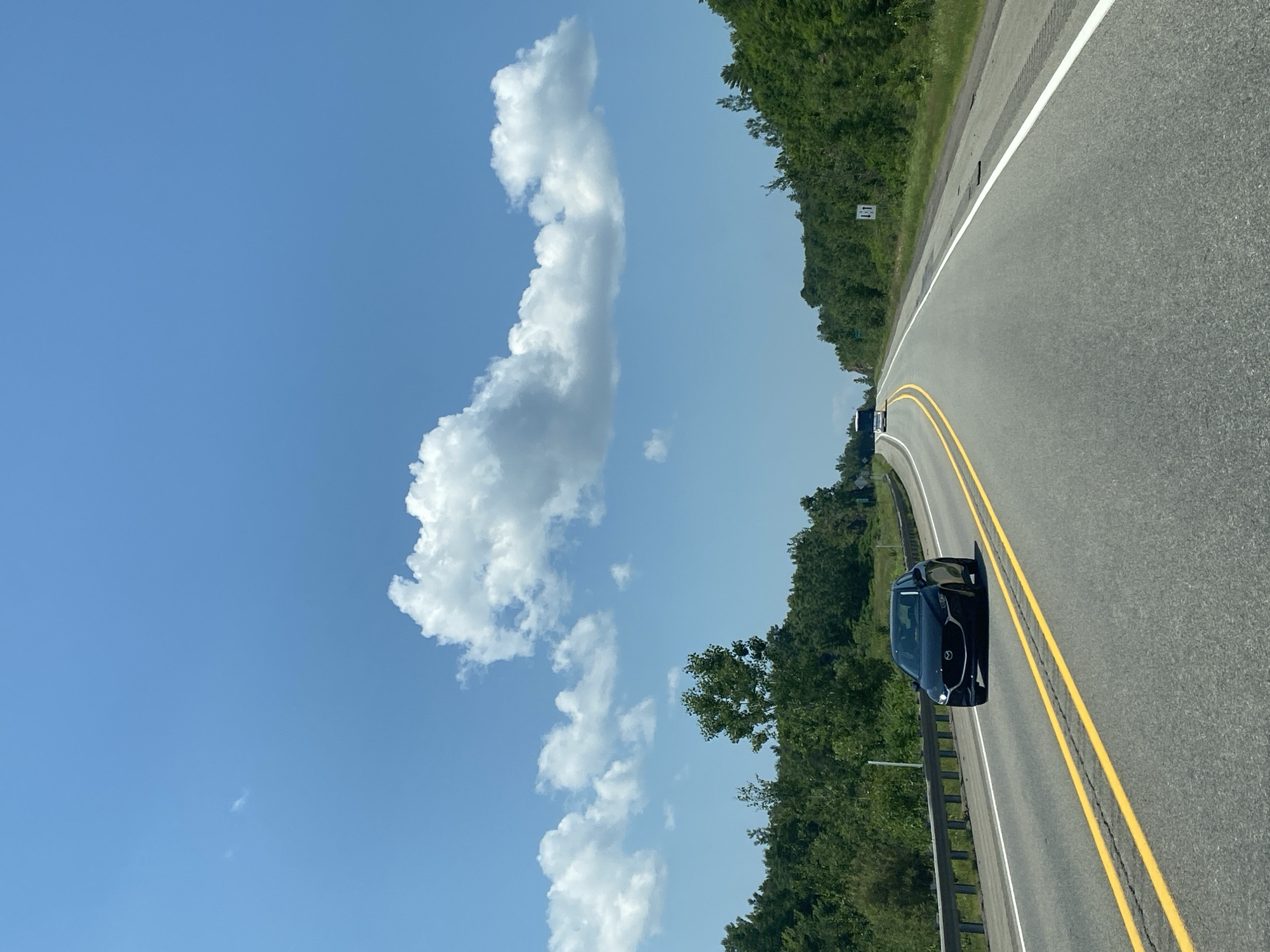 The image shows a paved highway in Pointe-au-Chêne, Canada, on a clear day with a bright blue sky and scattered white clouds. The road has two lanes in each direction, separated by double yellow lines. A black car is driving in the right lane, and a white bus is visible in the distance on the left lane. Lush green trees line both sides of the highway. A metal guardrail is present on the left side of the road, separating it from a grassy shoulder. The overall scene depicts a typical highway landscape during daytime travel.