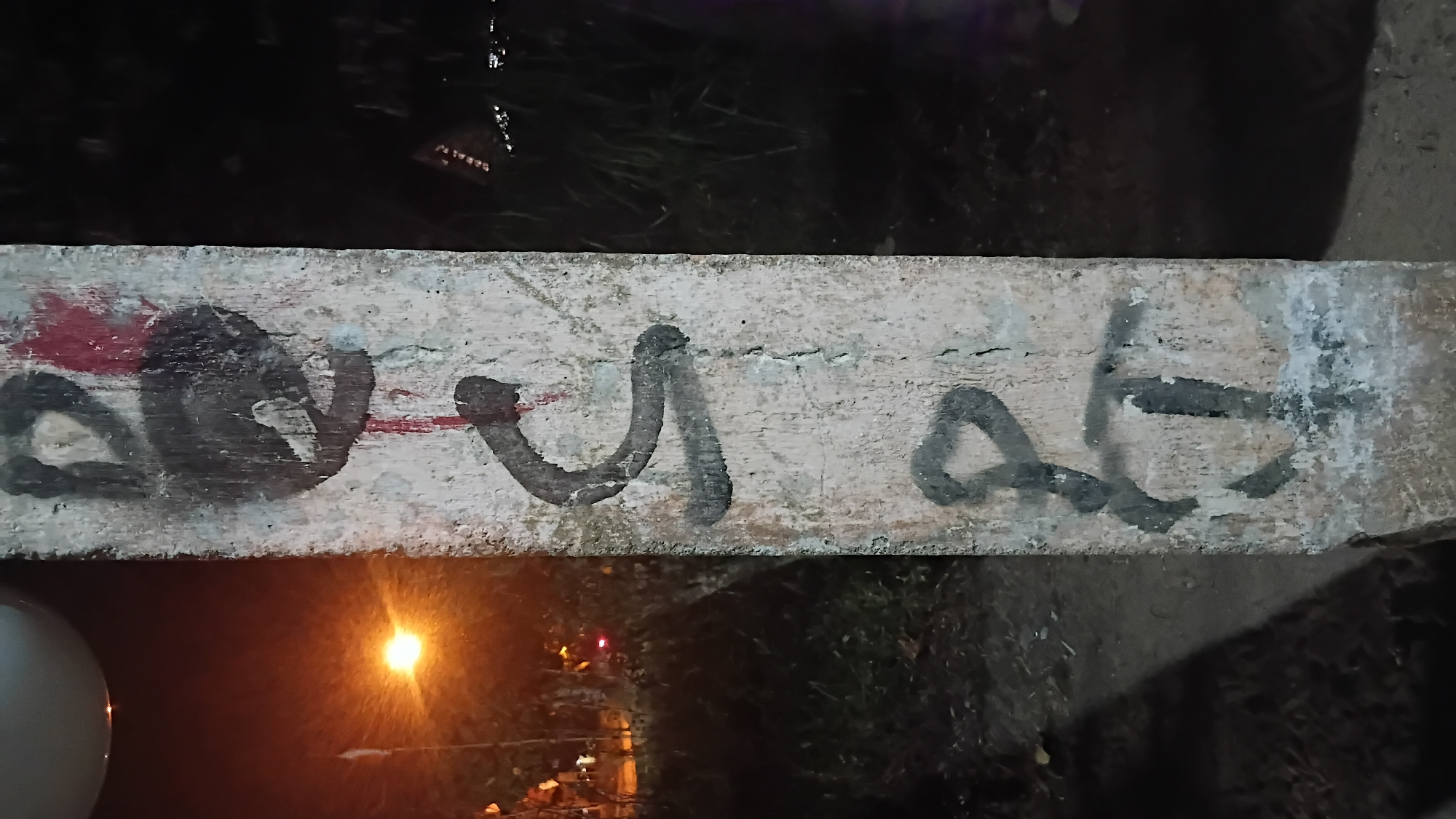 The image shows a concrete pillar with graffiti at night. The pillar is located in the city of Prabumulih, Indonesia. The graffiti consists of black and red markings, with some of the black markings appearing to be letters or symbols. In the background, there are trees and a street light, indicating an outdoor urban setting. The lighting suggests it is nighttime or dusk. The pillar itself appears weathered and aged.