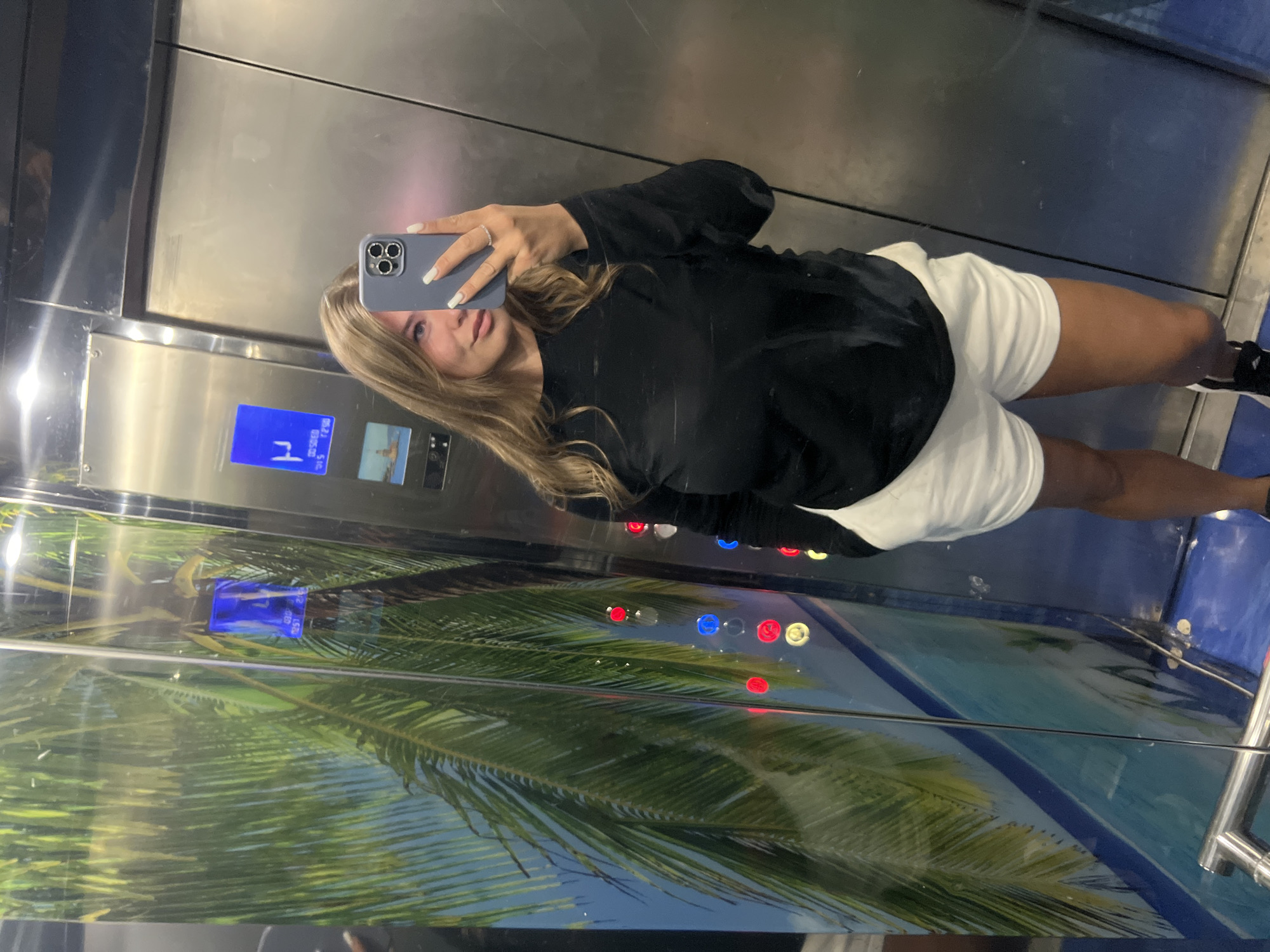 A woman stands in an elevator, holding a smartphone to take a selfie. She is wearing a black long-sleeved top and white shorts with black and white sneakers. The elevator has mirrored walls adorned with a palm tree design. The control panel of the elevator is visible. The elevator's destination display is also visible. The location is likely in Budva, Montenegro, though this cannot be definitively confirmed from the image alone.