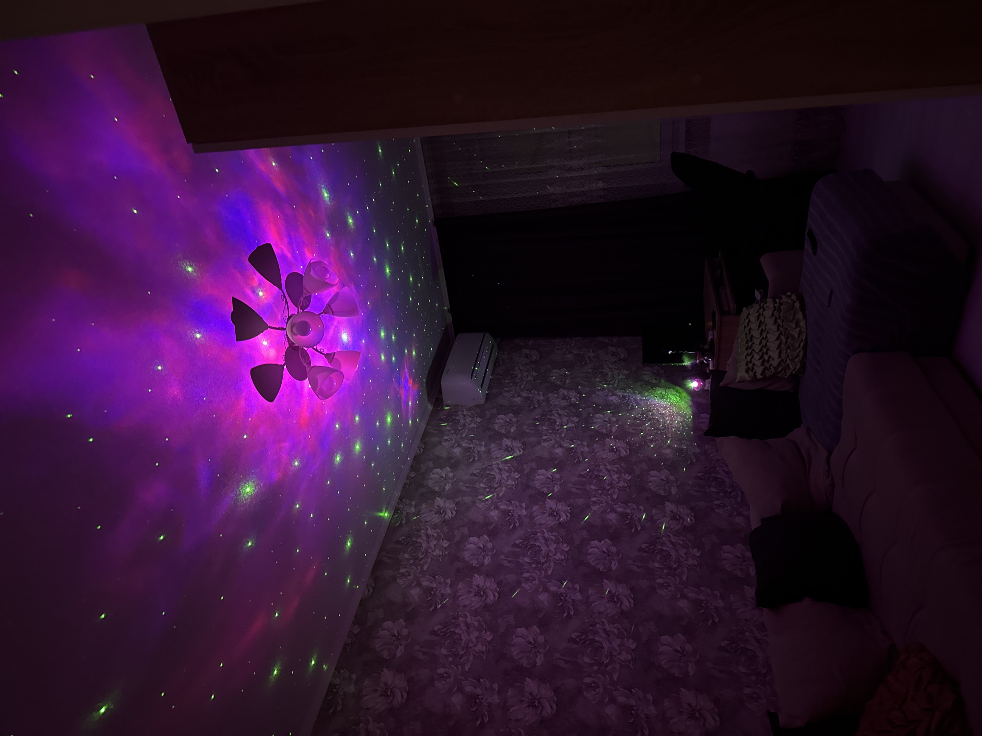 The image depicts an interior space with an elaborate lighting setup, featuring a ceiling-mounted fixture projecting purple and green star-like patterns onto the wall and possibly the floor. The wall on the left is lit up with pink, red, and purple hues. The floor is covered in a floral patterned carpet in a mix of purple and white. In the background, on the right, there's a couch with pillows and a large travel suitcase. A ceiling beam crosses the upper part of the image, obscuring the view of the ceiling in that area. There appears to be a window partially blocked by a dark object or material. The setting is an indoor space, possibly a room.