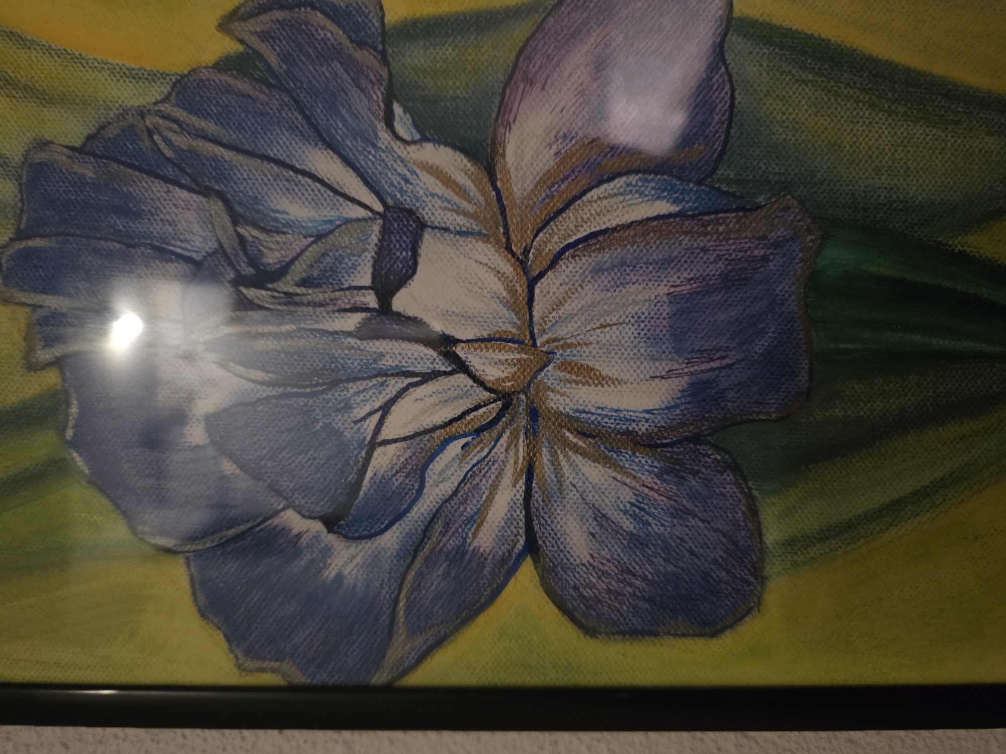 The image shows a framed painting of a blue and purple flower with green and yellow background. The painting is close-up, filling most of the frame. There are reflections visible on the glass covering the painting. The frame is black and the bottom edge of the frame is visible.