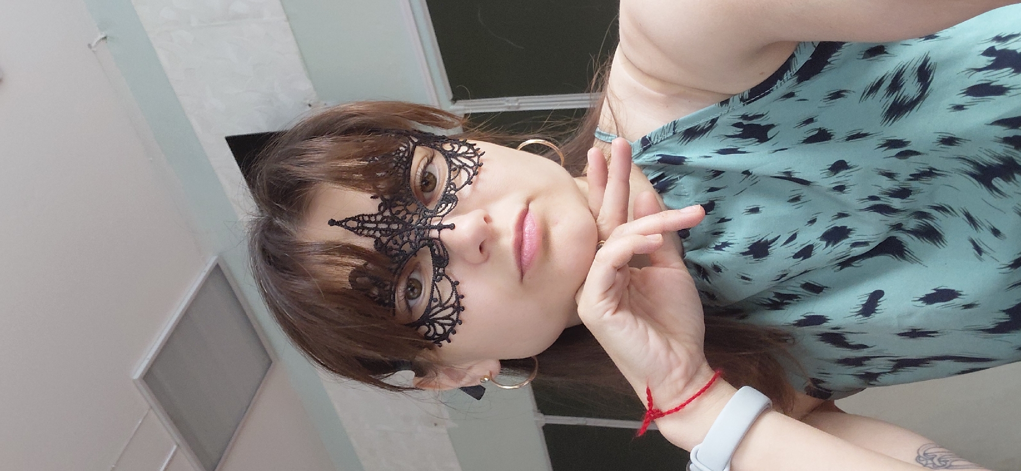 A young woman with brown hair and bangs wears a black lace masquerade mask that covers her eyes and the bridge of her nose. She is looking directly at the camera with a neutral expression, her chin resting on her left hand. She has light brown eyes and her lips are slightly parted. She is wearing large gold hoop earrings, and a thin red string bracelet is tied around her left wrist, above a white smartwatch. Her sleeveless top is a light teal color with a dark blue abstract pattern. The background appears to be an indoor setting with a light-colored ceiling and a faint blue wall. The image is a selfie taken from a slightly high angle.