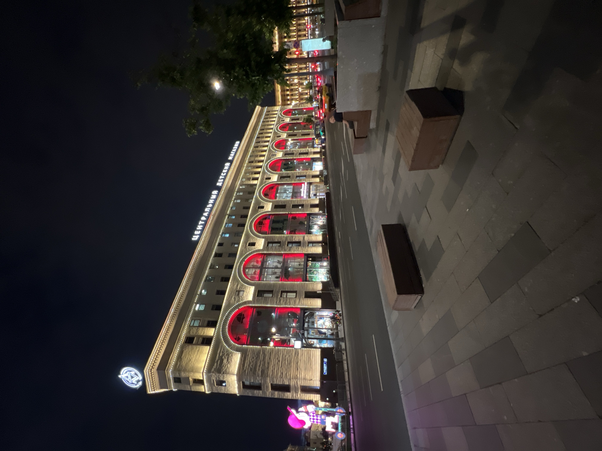 The image depicts a wide-angle, low-angle shot of the Central Children's Store (Tsentralny Detsky Magazin) in Moscow, Russia, at night. The building is illuminated with warm yellow lights along its facade and bright red lights within its arched windows. A prominent neon sign at the top of the building reads "ЦЕНТРАЛЬНЫЙ ДЕТСКИЙ МАГАЗИН". To the left of the building, a large, illuminated figure of a child on a scooter stands on the sidewalk. The street in front of the store is empty of pedestrians, but there are a few cars visible on the road, including a yellow taxi. Two wooden benches are placed on the sidewalk in the foreground. The sky is dark, indicating it is nighttime.