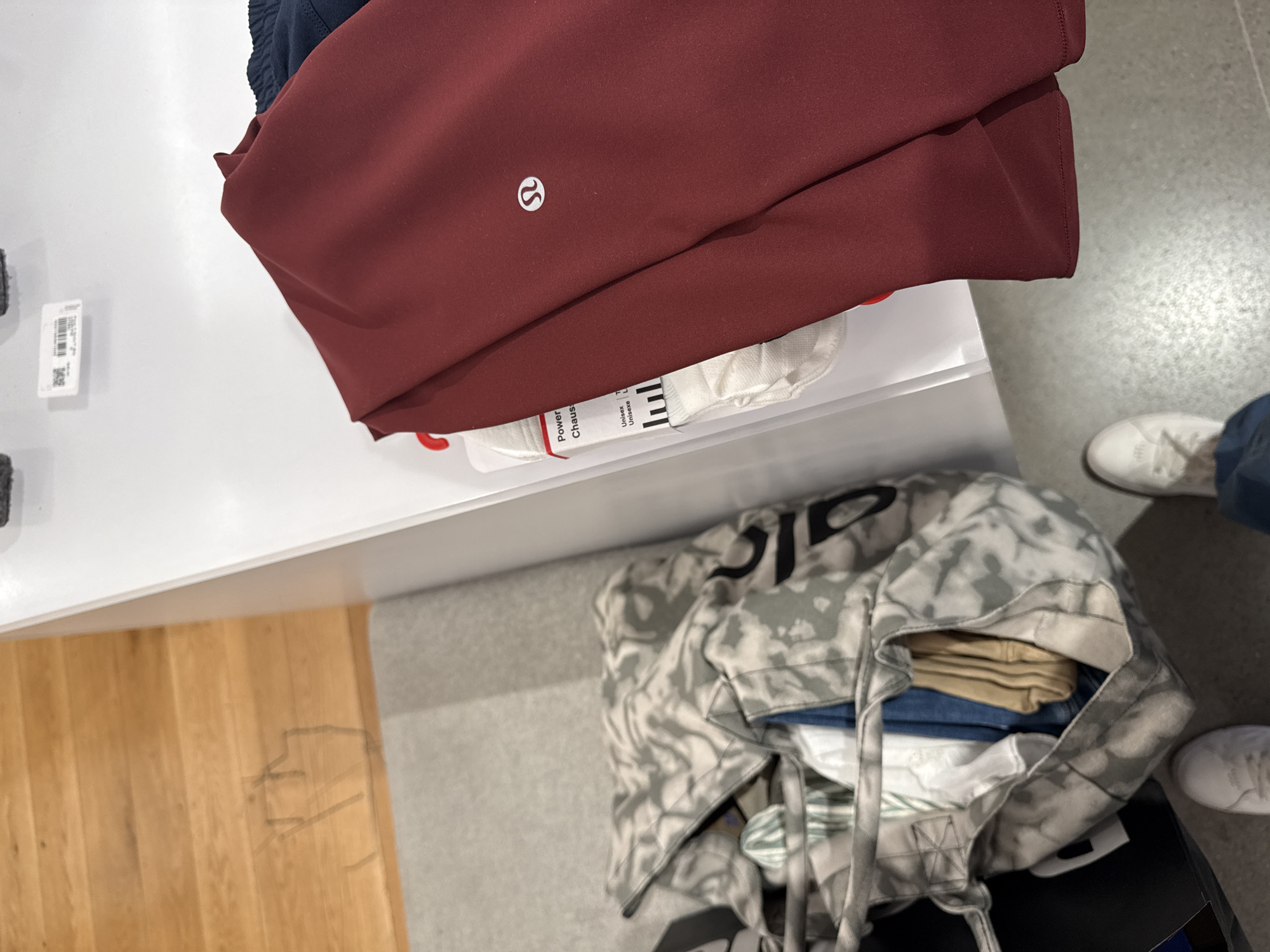 The image shows a display table in what appears to be a retail environment. On the table is a folded burgundy garment with a visible logo, possibly clothing. A white bag containing what looks like clothing and other items sits near the table. Part of a wood floor is visible on one side, while the other side reveals a grey floor. Two white shoes and a person's leg are also within the frame. The image does not provide definitive information about San Antonio or any specific location.
