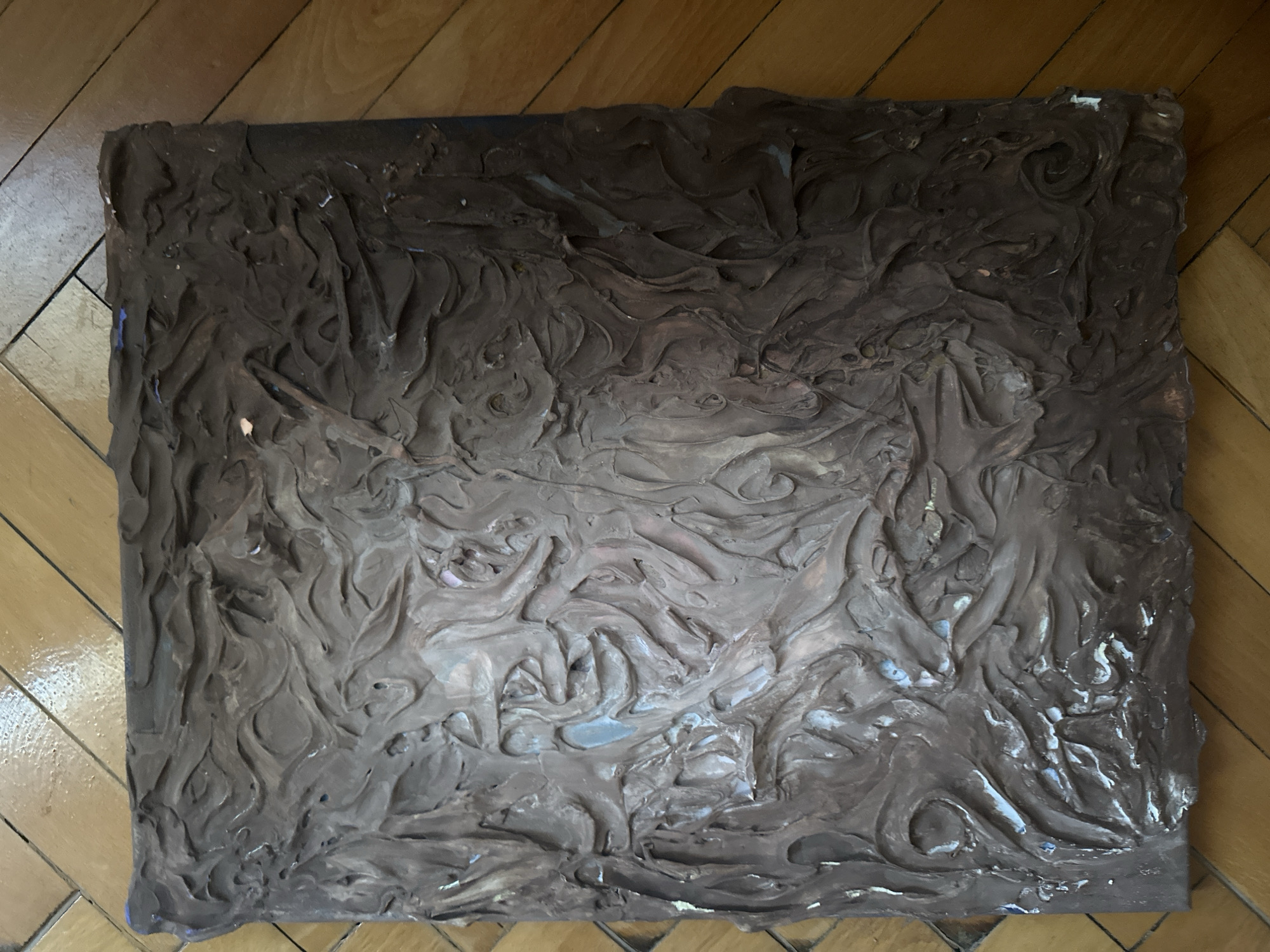 The image shows a square, textured artwork, possibly a relief or sculpture, with a dark brown, earthy tone. The surface is heavily impastoed with swirling, organic patterns that create deep shadows and highlights. The edges of the artwork appear somewhat rough and uneven, with some exposed lighter material underneath and small flecks of blue and pink paint visible on the upper edges. The artwork is placed on a polished wooden floor with a chevron pattern, characteristic of older European architecture. The lighting suggests an indoor setting, likely a home or studio. There is no visible indication of the city of Plovdiv, Bulgaria, or any people in the image.