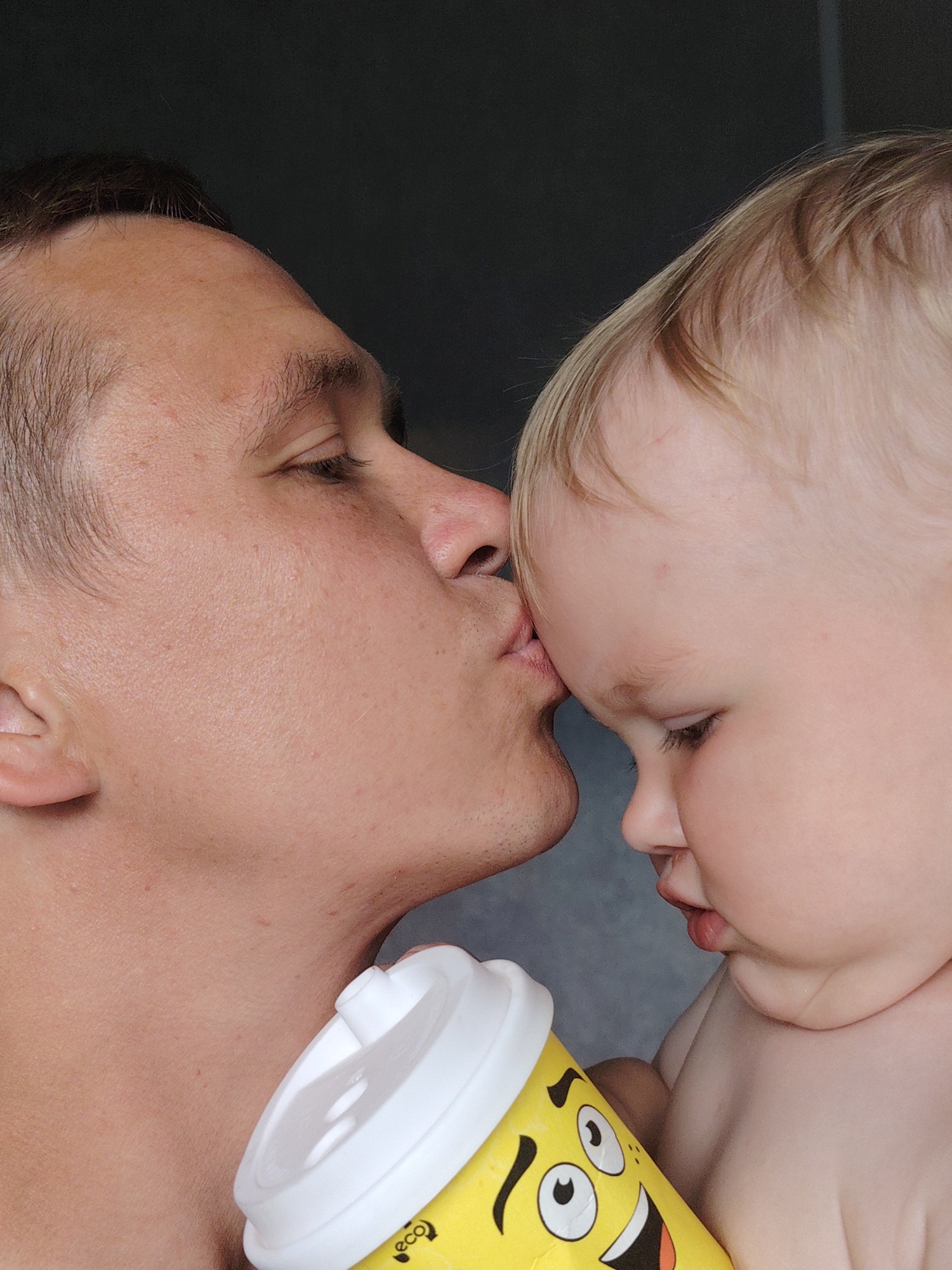 In a close-up, profile view, a man gently kisses the forehead of a baby. The man's head is on the left side of the frame, and he is looking down at the baby. His eyes are closed, and his lips are pressed against the baby's skin. The baby's head is on the right side of the frame, and the baby is looking down and away from the man. A portion of a yellow cup with a smiley face graphic is visible in the lower portion of the frame, held by the baby. The background is a dark, solid color, suggesting an indoor setting. There are no discernible landmarks or environmental features to indicate a specific location in Chaikovsky, Russia.
