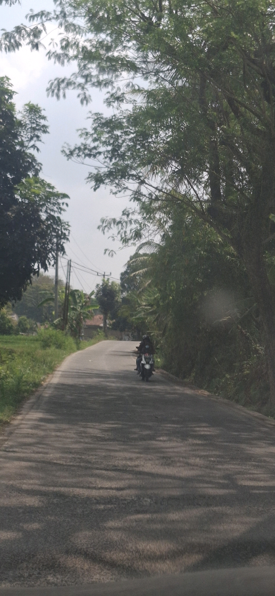 A motorcycle with two occupants is traveling on a paved road in Cibinong Hilir, Indonesia. The road is flanked by lush green vegetation, including trees and foliage, with some scattered buildings visible in the background. The sky is overcast, and shadows from the trees are cast across the road. The scene depicts a rural or semi-rural environment.