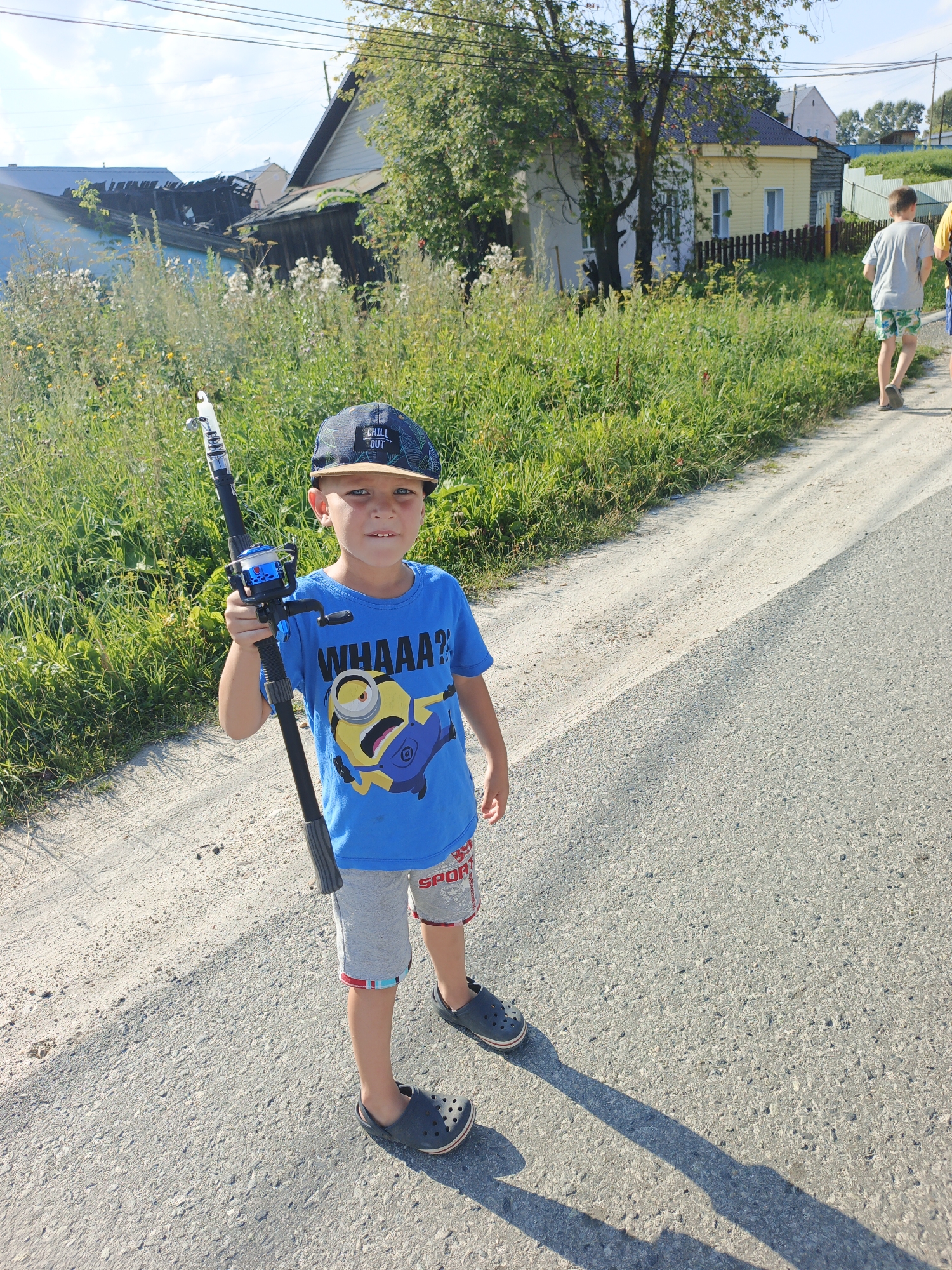 A young boy, wearing a blue t-shirt with a cartoon character, gray shorts, a dark baseball cap, and dark clogs, stands on a dirt road in Medvedyovka, Russia. He is holding a fishing rod. In the background, there are houses, green grass, and trees under a clear blue sky. Another person, a young boy in a grey t-shirt and colorful shorts, is walking away from the camera down the road.