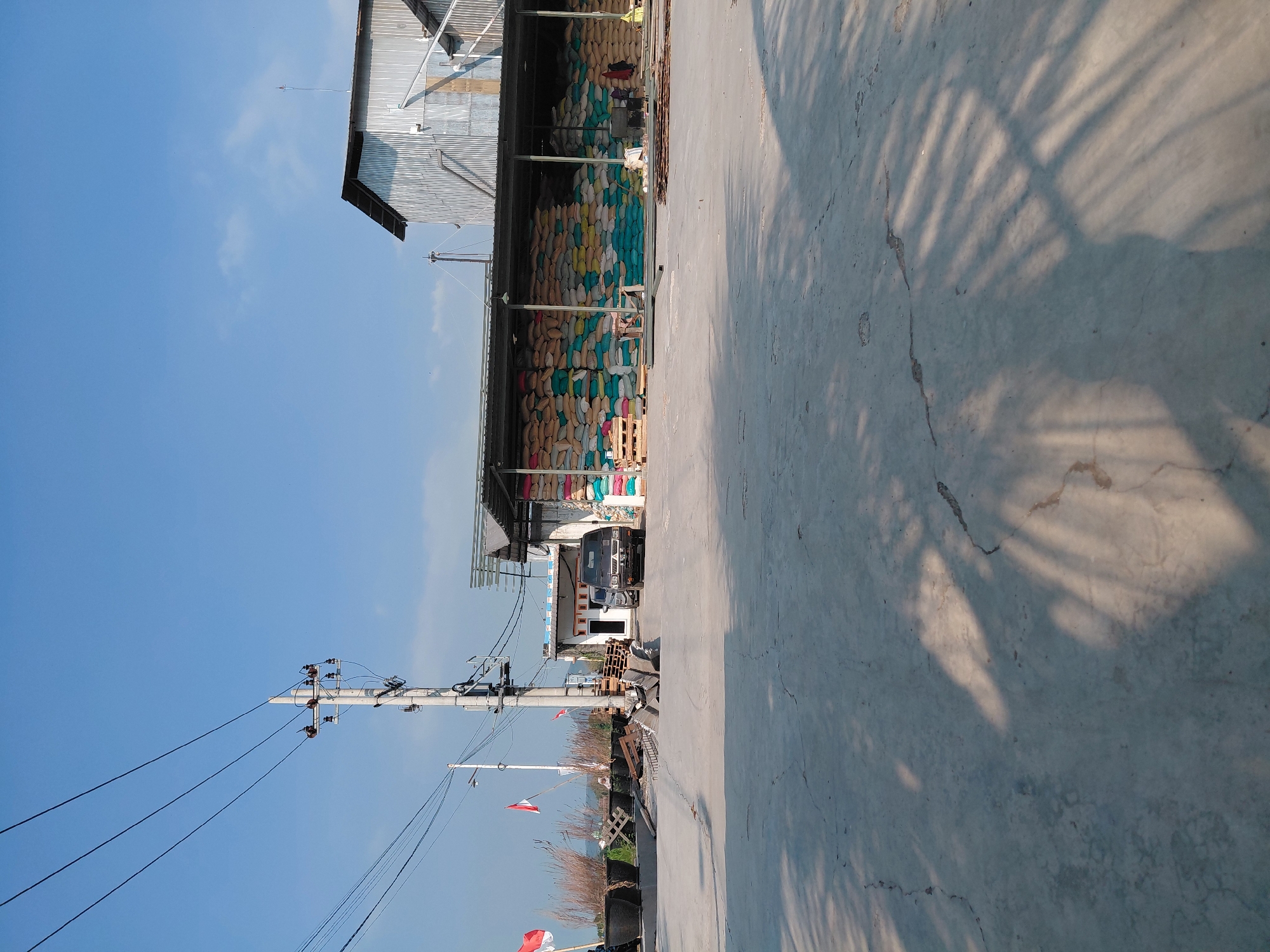 This is a horizontal, eye-level, medium-shot photograph taken outdoors in Alas Tengah, Indonesia. The image captures a scene outside a warehouse or storage facility on a clear, sunny day with a bright blue sky. In the foreground, a cracked concrete surface with shadows of palm leaves is visible. In the middle ground, there is a paved area leading to a building with an open-sided roof. Large stacks of colorful sacks, likely containing agricultural products such as rice or fertilizer, are piled high under the roof. Two cars are parked in front of the building, and a utility pole with electrical wires stands prominently to the left. Red and white Indonesian flags are displayed on flagpoles near the left side of the image, and large black planters are also present. In the background, a low-rise building with a corrugated metal roof is visible, along with more stacks of sacks. The overall scene suggests an active industrial or agricultural storage area.