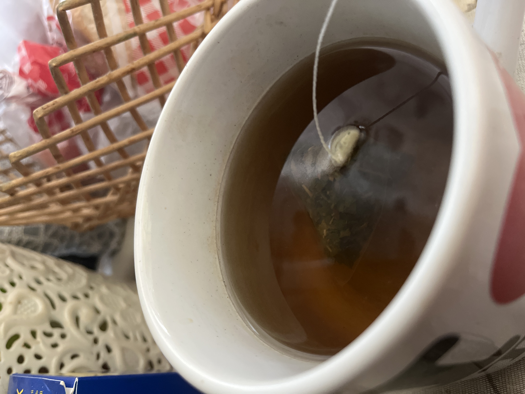 An overhead, medium shot captures a white mug filled with amber tea and a tea bag submerged within. The mug has a portion of a heart design visible on its exterior. In the background, a woven basket with a red and white checkered cloth and what appears to be bread is partially visible. To the left, a portion of a blue box with some text in Cyrillic is also in view. The lighting suggests an indoor setting. There are no people or specific landmarks to indicate a location in Minsk, Belarus.