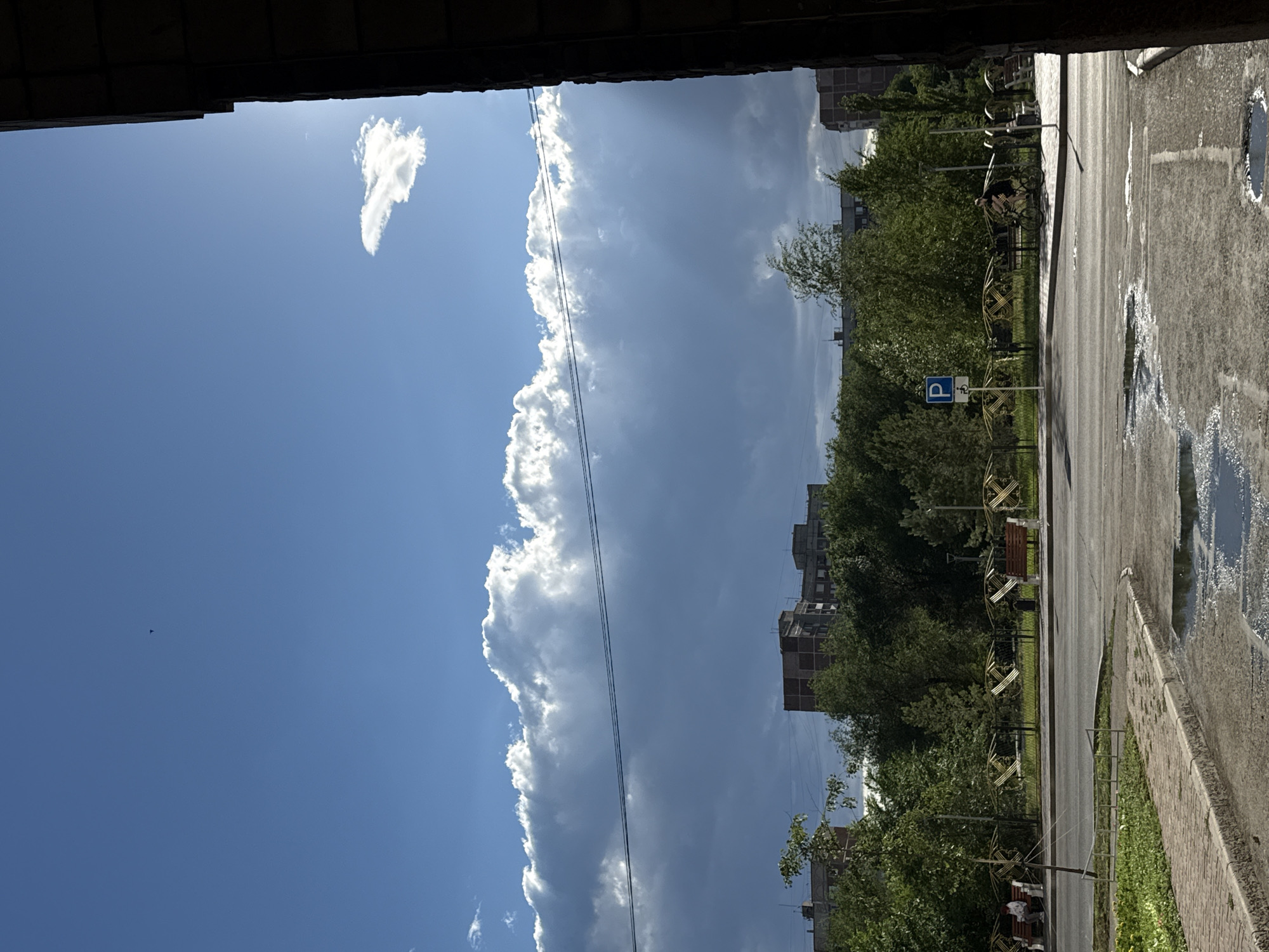 The image, taken from an elevated vantage point, likely a balcony or window, depicts an urban scene in Pavlodar, Kazakhstan. The sky dominates the left side of the frame, a vibrant blue dotted with fluffy white clouds and a larger, dramatic cloud formation stretching across the mid-section. The sun is visible, casting rays through the clouds, suggesting a partly sunny day.
On the right, a paved area, possibly a street or sidewalk, is wet with puddles reflecting the sky, indicating recent rain. A brick curb separates the pavement from a grassy area. Beyond this, a line of trees with lush green foliage forms a natural barrier. In the background, several multi-story buildings, some appearing to be under construction, rise above the trees.
Further into the scene, a road is visible, with a parked bicycle and a person sitting on a bench under a tree. A blue parking sign with a white "P" is posted. The overall impression is of a calm, everyday moment in a residential urban area, possibly mid-afternoon given the quality of light. The weather appears to be clearing after a period of rain. No discernible text is visible besides the "P" on the parking sign.