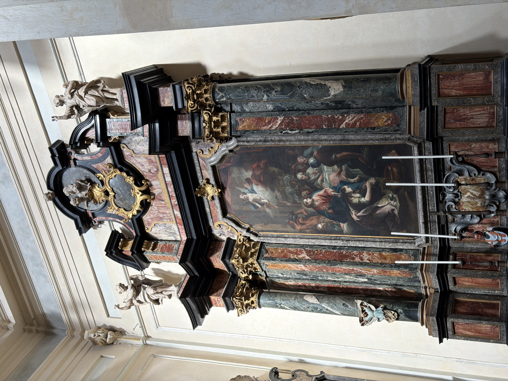 The image shows an ornate baroque-style altar in a church in Milan, Italy. The altar features marble columns, intricate gold leaf ornamentation, and sculptures of angels. The central painting depicts a scene with numerous figures, possibly a religious narrative. The overall aesthetic is highly decorative and characteristic of baroque art.