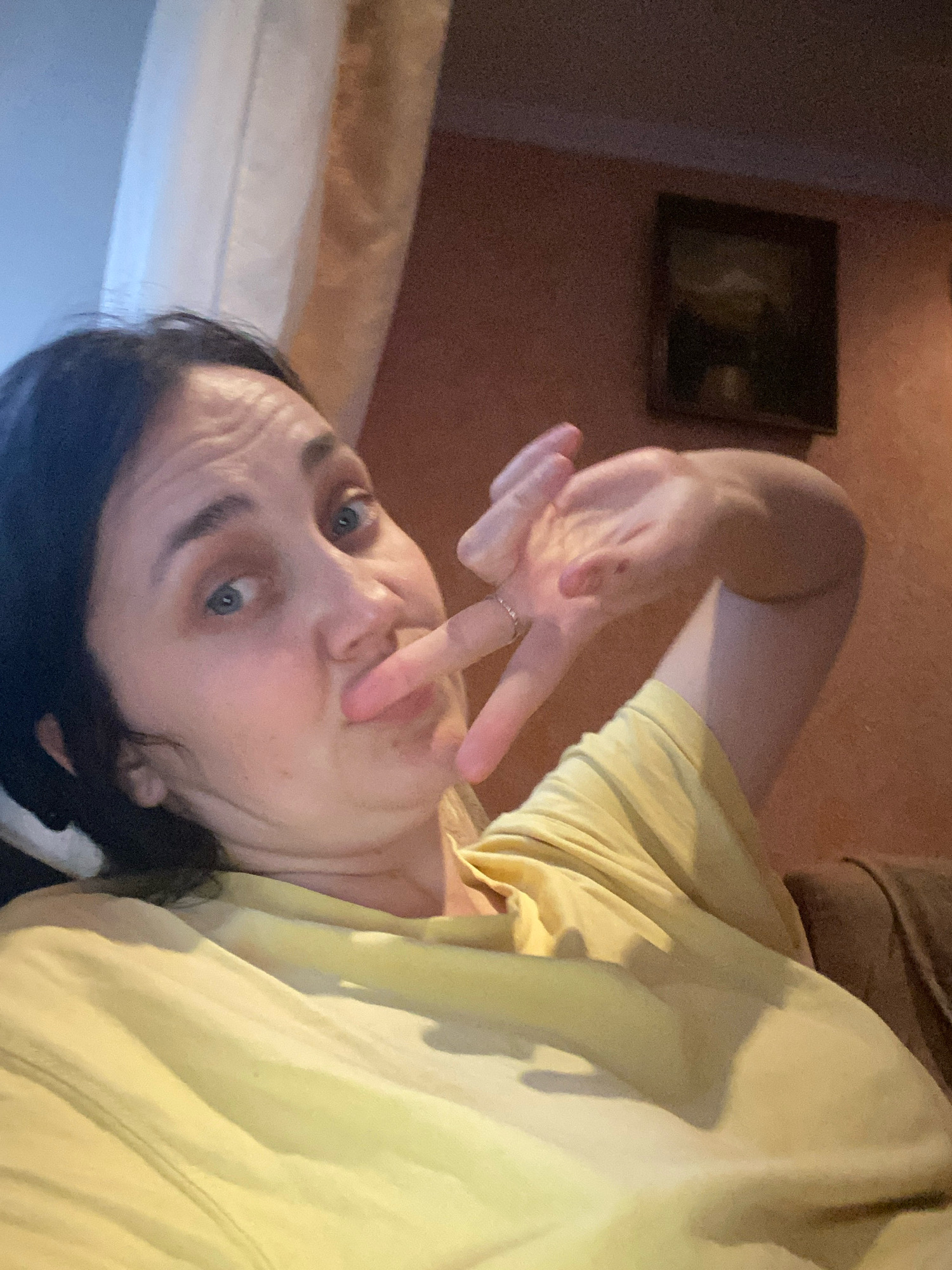 A woman is lying down indoors, possibly on a couch, wearing a yellow t-shirt. She is looking at the camera and making a peace sign with her right hand, which is near her face. Her left hand is also visible, resting on her chest. There is a framed picture on the wall in the background, which appears to be a landscape painting. A light-colored curtain is visible on the left side of the image. The lighting suggests it is daytime, with some natural light coming from the left.