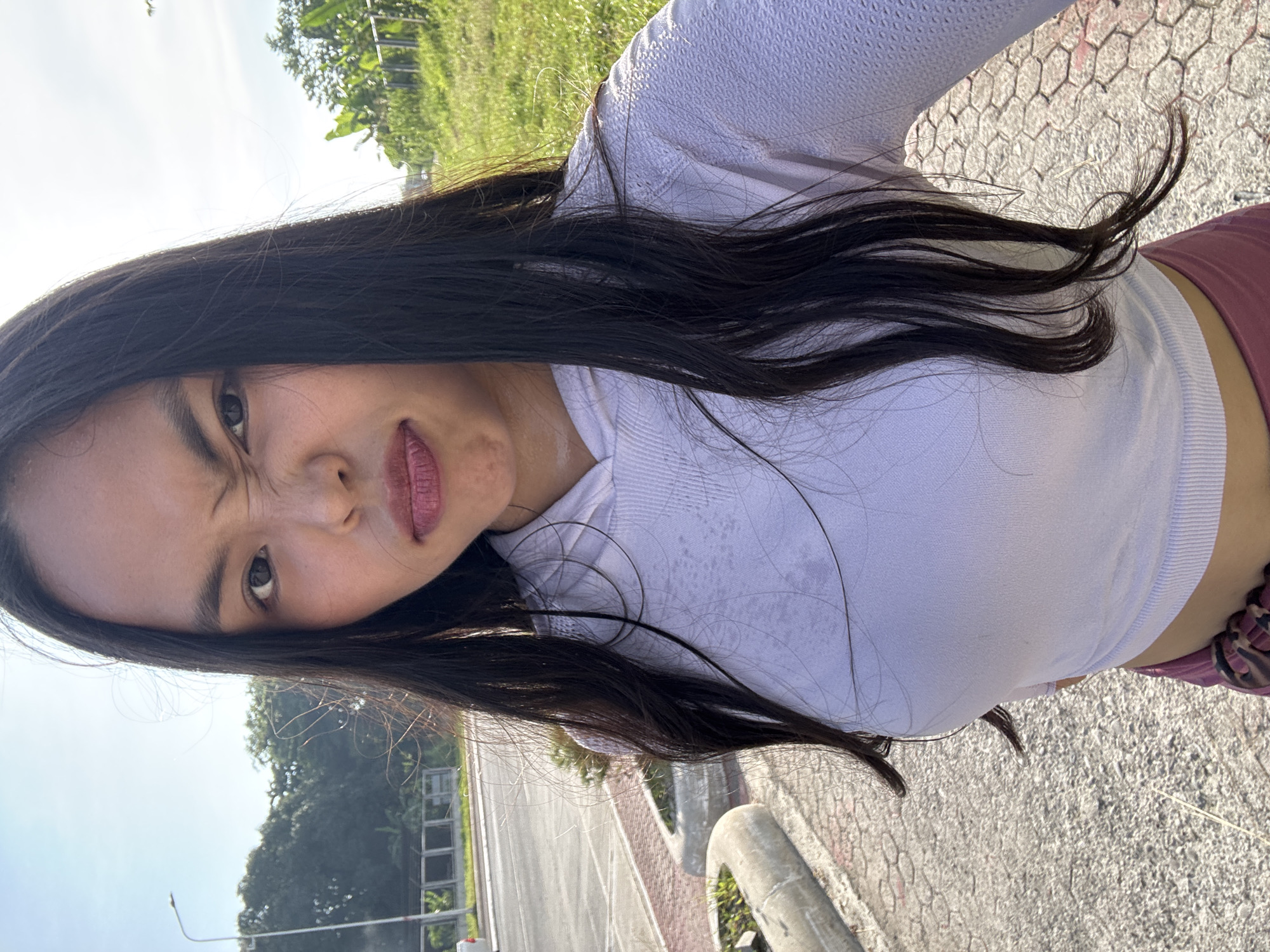 A young woman with long, dark hair is taking a selfie outdoors. She is wearing a white cropped hoodie and pink pants. She is outdoors with a street, sidewalk, and grassy area visible in the background. The sky is bright and sunny, suggesting it is daytime. The presence of palm trees and the overall appearance of the vegetation indicate a tropical climate, consistent with Davao City, Philippines. The background also shows a street lamp and a road, typical of an urban or suburban environment within the city.