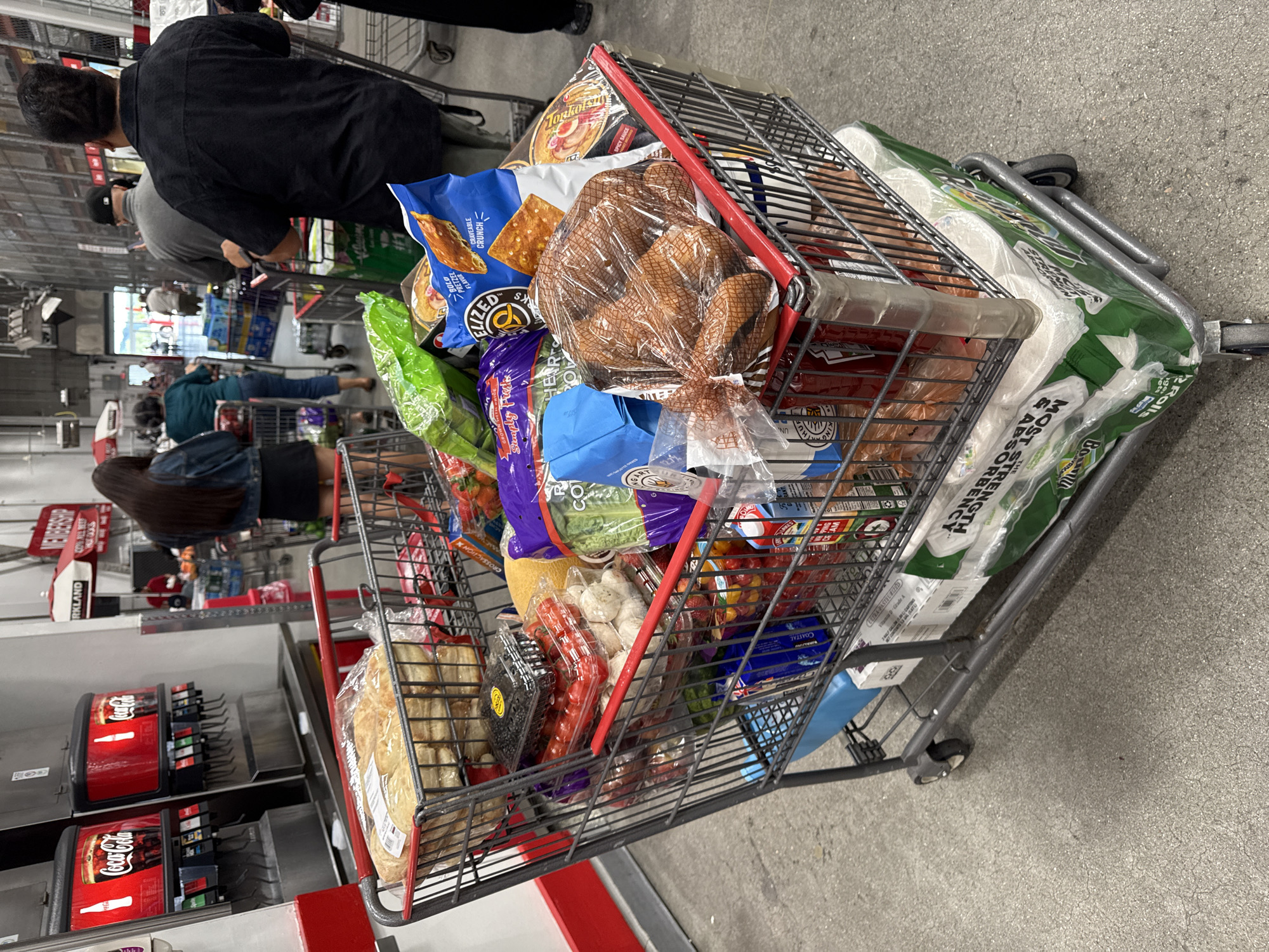 In the City of Yonkers, United States of America, a grocery shopping cart is filled with various items. The cart is positioned on a concrete floor, with a Coca-Cola soda fountain visible in the background. A woman in a denim jacket and black skirt is standing to the left of the cart, facing away from the camera. The cart contains a bag of bread, a container of avocados, a container of cherry tomatoes, a bag of pretzel crisps, a bag of pretzels, a package of hot dogs, a package of lettuce, and a package of toilet paper. Other shoppers and shopping carts are present in the background.