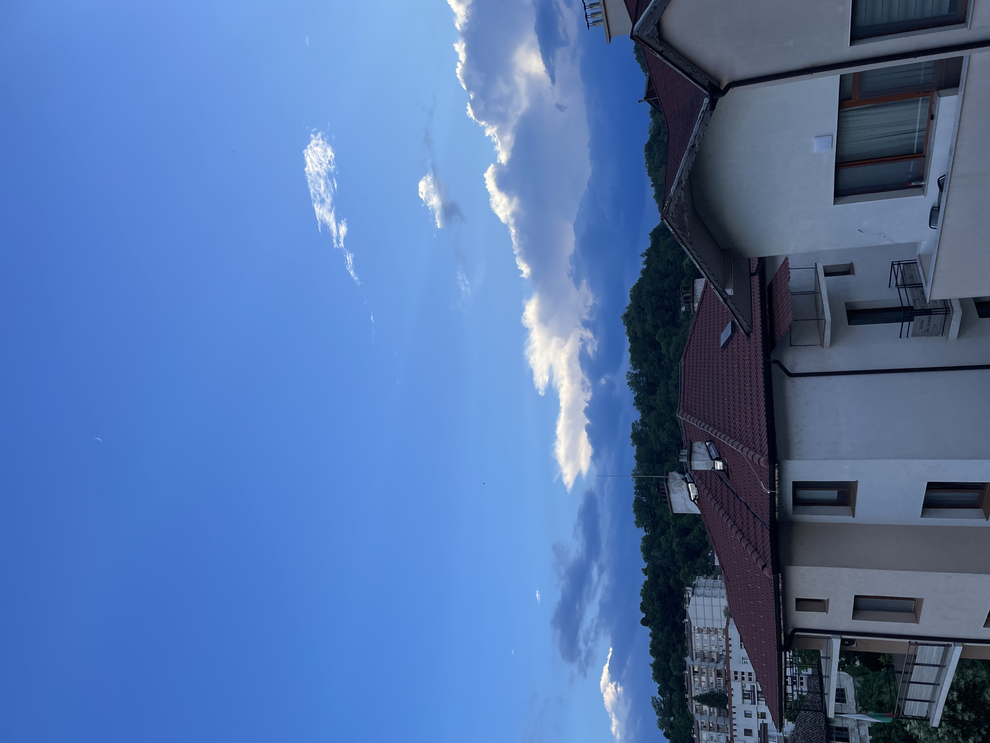 The image depicts a view of buildings and a forested hillside under a blue sky with scattered clouds. The buildings appear to be residential, with tiled roofs and multiple stories. Some have balconies with railings, and windows are visible. In the foreground, a white building with a reddish-brown tiled roof dominates the lower right of the frame. To the left, another building with a similar roof is partially visible. Further back, a block of taller, lighter-colored buildings is situated on the hillside. The hillside itself is covered in dense green trees. The sky is a vibrant blue, with some wispy white clouds and a crescent moon faintly visible in the upper left quadrant. The scene suggests an urban environment with natural surroundings. The image seems to be taken from a slightly elevated perspective, looking across rooftops and toward the hillside.