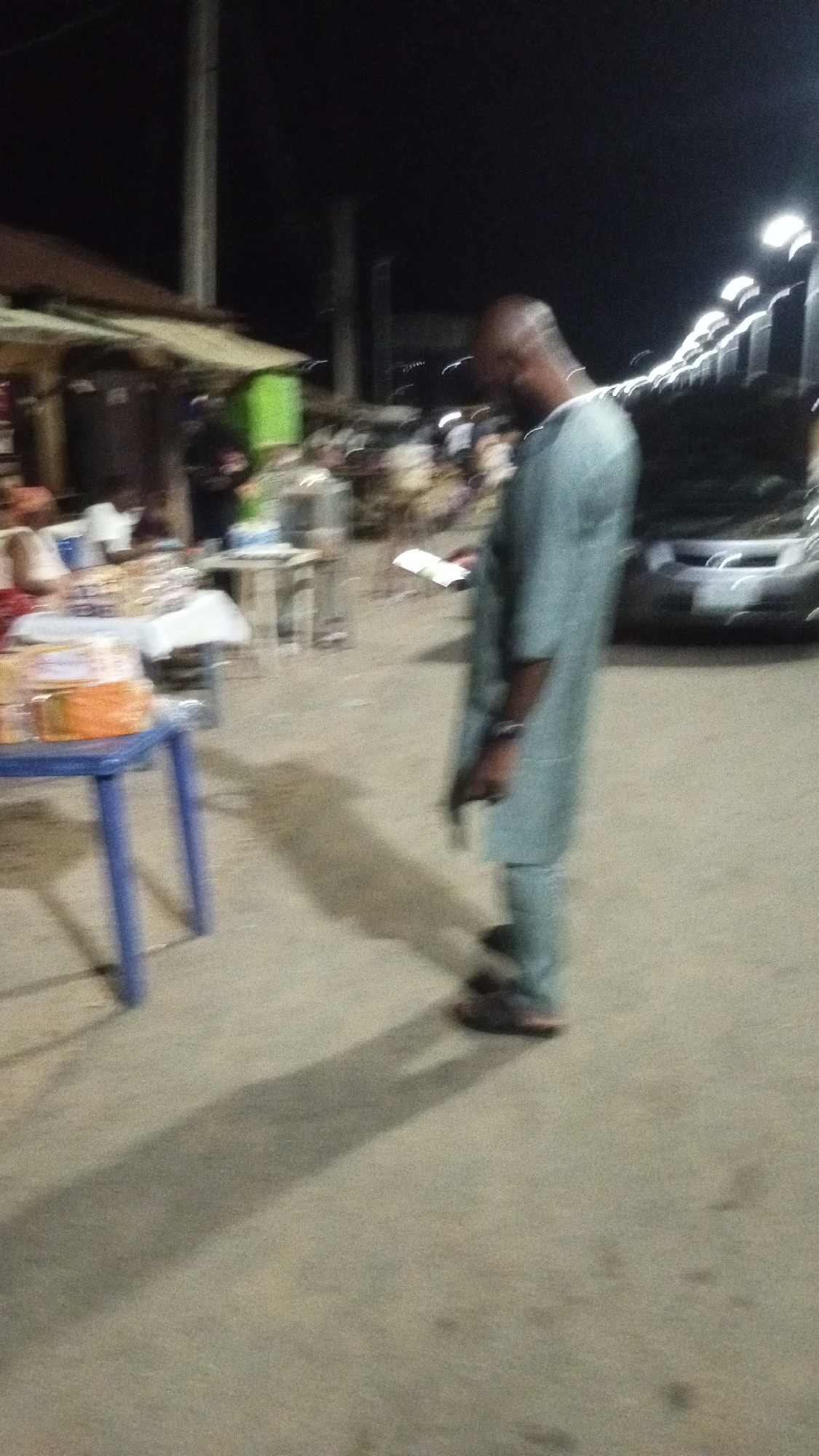 The image depicts a night scene in an outdoor market or street setting, likely in Adankolo, Nigeria. A bald man, wearing a light blue traditional tunic and trousers with dark sandals, stands slightly to the right of the frame, looking downwards or to his left. He holds a small, rectangular white object in his right hand.
The scene is illuminated by artificial lights, casting long, distinct shadows on the concrete or asphalt ground, including a prominent shadow of the man stretching into the foreground. To the left, several market stalls or makeshift structures are visible, with items, possibly packaged food, displayed on tables, one of which is a blue plastic table. Other blurred figures and tables can be seen in the mid-ground. In the far background to the right, part of a light-colored car is visible, along with a row of streetlights emitting a bright glow. The image exhibits blurriness throughout, suggesting low-light conditions or motion during capture. No visible text can be clearly read.