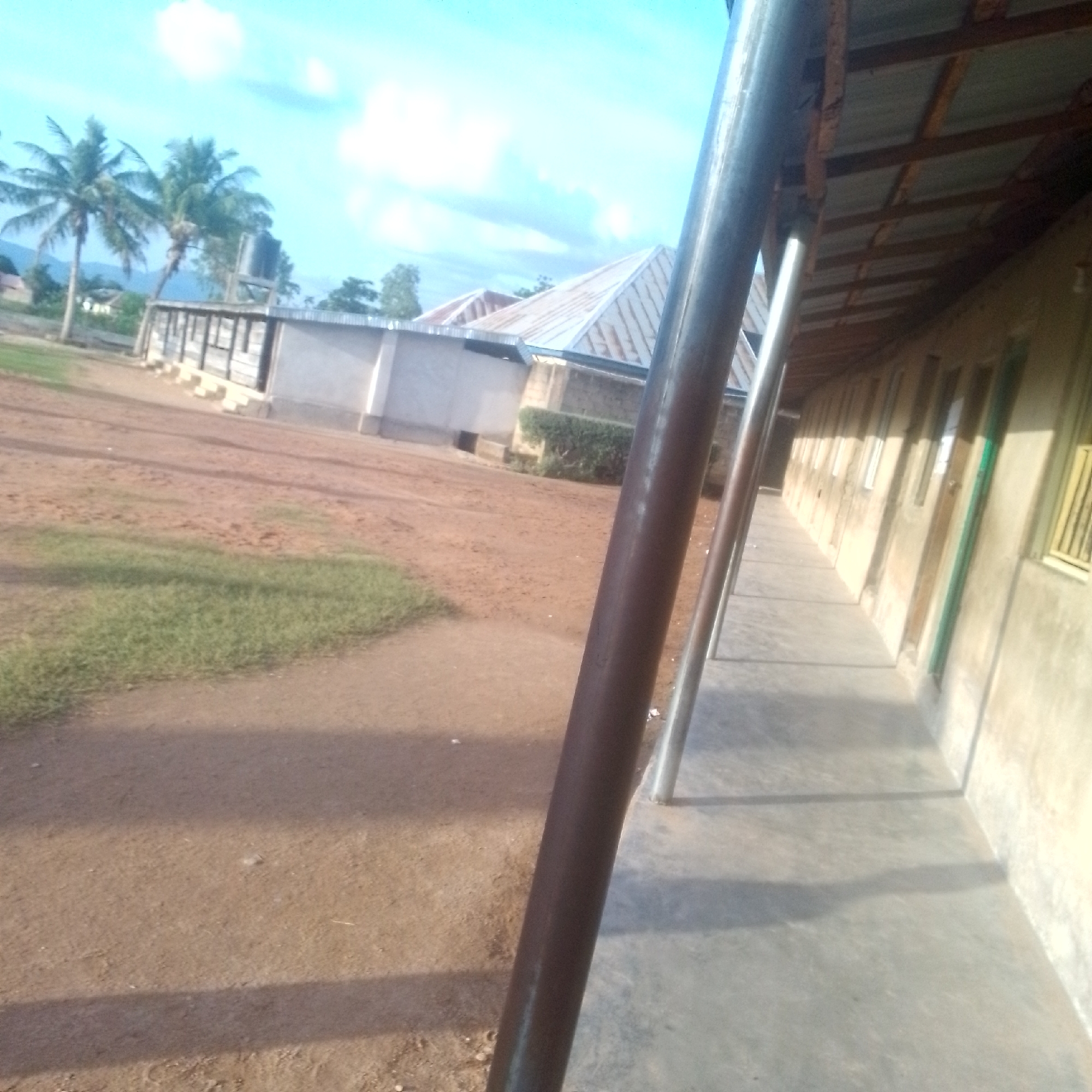 This media file depicts an outdoor scene on a bright, sunny day, likely in Jalingo, Nigeria. The foreground is dominated by a dirt pathway and a patch of green grass. In the background, there are several buildings with corrugated metal roofs. One building appears to be a single-story structure with a water tank on top, and it is flanked by palm trees. Another building, seen further back, also has a distinctive metal roof.
Along the right side of the image, a long, narrow building stretches into the distance. This building has a covered walkway or veranda with a concrete floor, supported by metal posts. The wall of this building is made of rough-textured plaster, and it features a series of windows and doors. The windows have barred coverings, and some doors are painted green. The sunlight casts long shadows across the walkway and the ground, indicating it is likely morning or late afternoon. The sky is a clear blue with scattered white clouds, suggesting fair weather.
There are no people visible in the image, and no text is present. The overall impression is one of a rural or semi-rural setting, possibly a school or a residential compound, with modest architecture typical of the region. The scene is peaceful and unremarkable, showcasing a slice of everyday life in Jalingo.