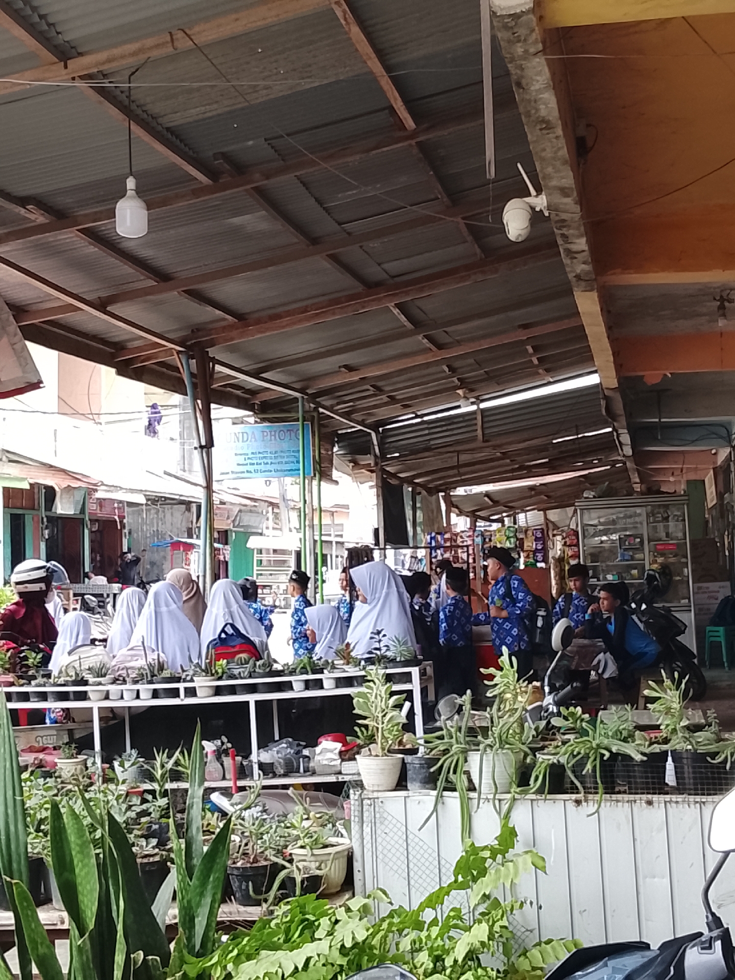 The image depicts a lively market or street stall scene in Lhokseumawe, Indonesia, characterized by a prominent display of potted plants in the foreground and midground. Various plant species, including succulents, snake plants, and other leafy greens, are neatly arranged on white multi-tiered shelving units.
Numerous individuals are present, many appearing to be students. Girls are seen wearing white headscarves (hijabs) over their uniforms, while boys wear distinctive blue patterned shirts, suggesting they are in school attire. They are gathered around the plant stalls, some seemingly browsing or conversing, contributing to a casual and interactive atmosphere.
The area is covered by a corrugated metal roof supported by a network of wooden and metal beams, providing shade. A single white LED light bulb hangs from the roof structure. On the right, a white dome-shaped CCTV camera is mounted on a concrete pillar. In the background, other storefronts are visible, including one with a blue sign prominently displaying "NDA PHOTO." Further to the right, a shop interior with shelves stocked with various items can be seen, and a motorcycle is partially visible parked nearby.
The scene is illuminated by natural daylight, indicating it is daytime. The overall setting is bustling and commercial, reflecting daily life in a local marketplace.