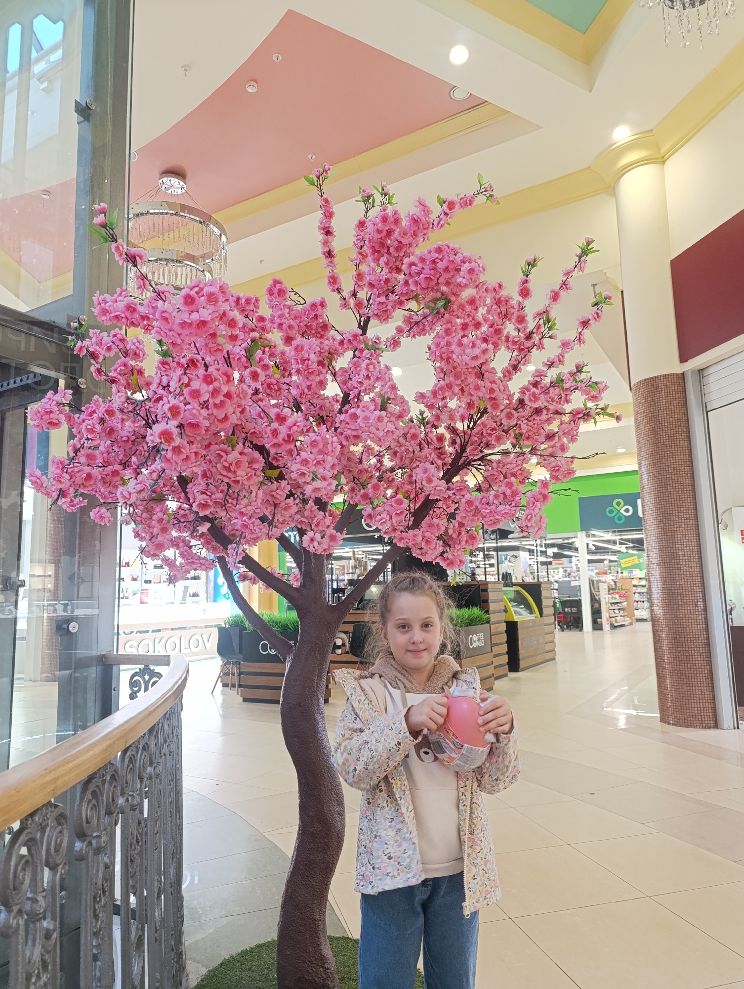A young girl stands in a shopping mall next to a decorative tree with pink blossoms. The girl is wearing a light-colored jacket with a floral pattern, a beige sweatshirt, and blue jeans. She holds a pink, round object in her hands. The tree, placed on a small patch of artificial grass, is positioned near a metal railing and a glass storefront. The mall's interior is visible in the background, with various shops and a food service area. The ceiling has a color scheme of pink, beige, and light blue.