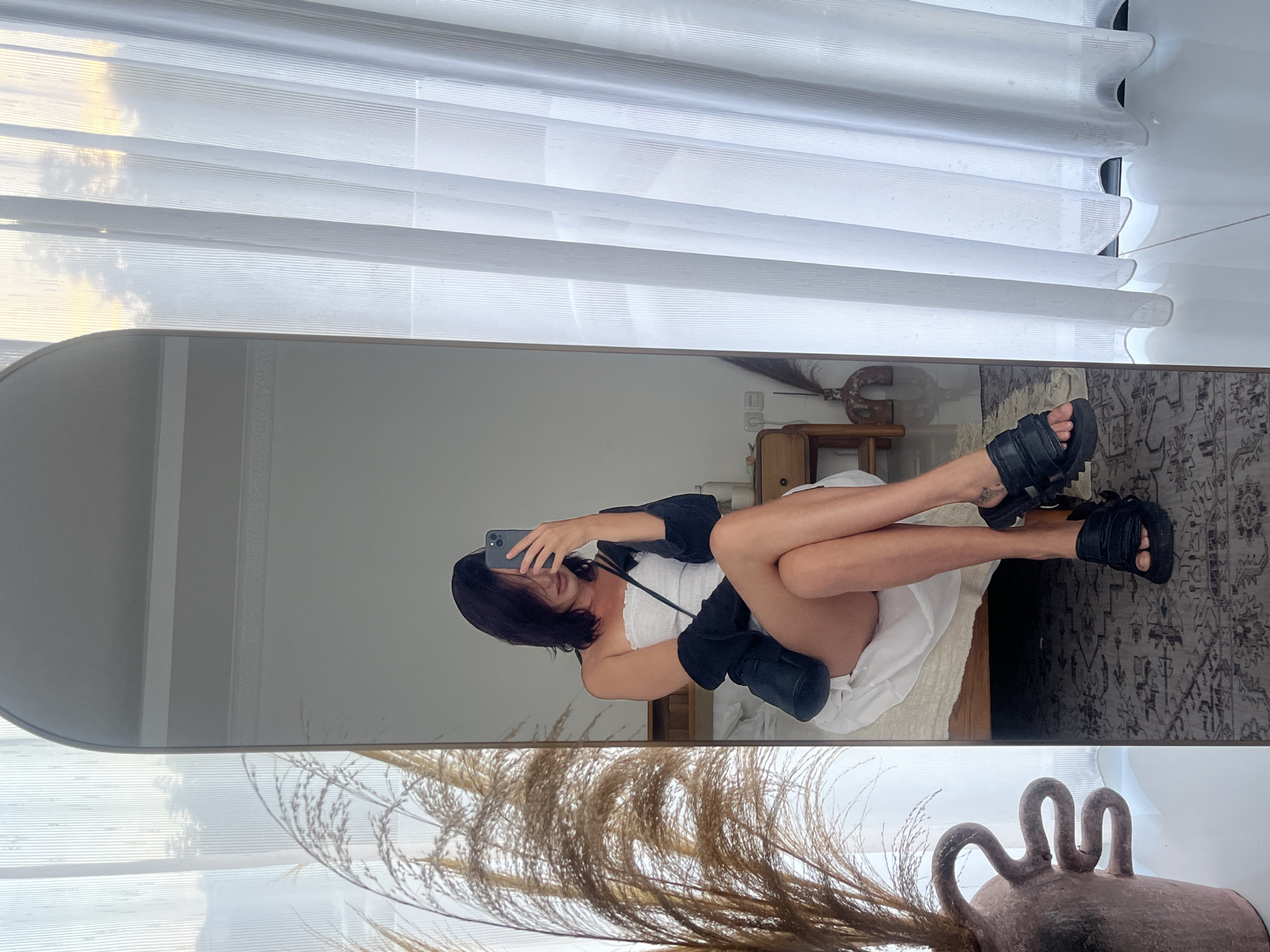 A stylish young woman is captured in a mirror selfie, reclining on a bed in what appears to be a well-lit bedroom in Kuta Selatan, Indonesia. She is positioned diagonally in the large mirror, with her head towards the left and her legs crossed towards the right. She wears a white strapless top or dress, a black long-sleeved jacket, and chunky black sandals. Her phone is visible in her hand as she takes the photo.
The reflection in the mirror reveals a minimalist and cozy room setting. Behind the woman, a bed with white bedding is visible. To the right, a wooden bedside table with various items rests beside the bed. A patterned rug covers a portion of the floor. In the background of the reflection, white horizontal window blinds are partially open, allowing soft daylight and hints of a cloudy sky to filter through.
In the immediate foreground, outside the mirror's frame, dried pampas grass or similar decorative foliage is arranged, partly obscuring a rustic, dark ceramic pot with distinctive loop handles. The overall ambiance is relaxed and bright, suggesting a leisurely daytime moment in a tastefully decorated personal space. There is no visible text in the image.