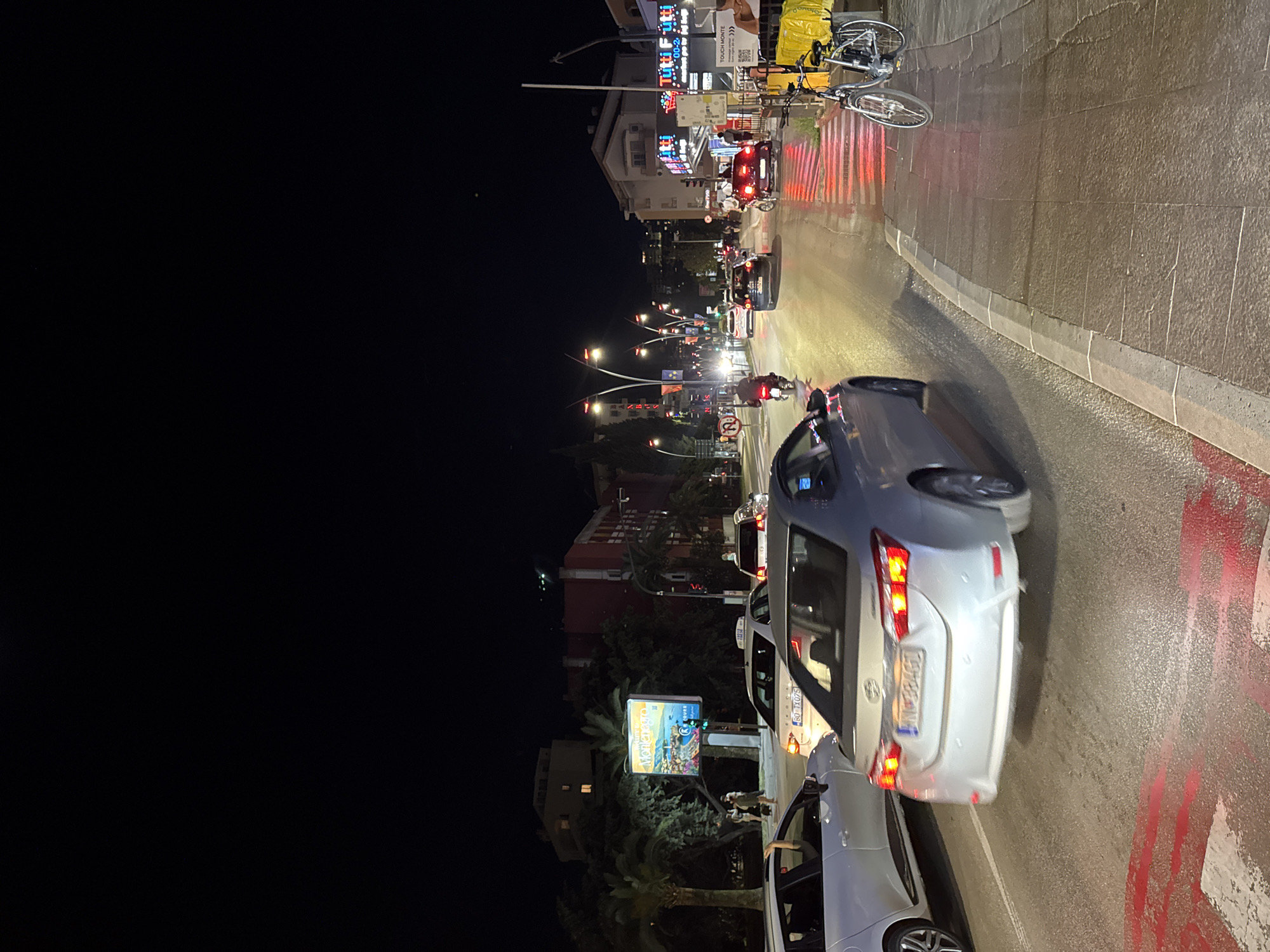 The image captures a nighttime street scene in Budva, Montenegro. A line of vehicles, including cars and possibly taxis, is visible on the right side of the image. Traffic lights and street lamps illuminate the road and surroundings. Billboards, buildings, and trees line the street. A bicycle is parked to the left of a store front that has illuminated signs. Pedestrians are not clearly visible. The road surface appears wet, reflecting the lights. The lighting condition is dark, with artificial lights creating a bright contrast.