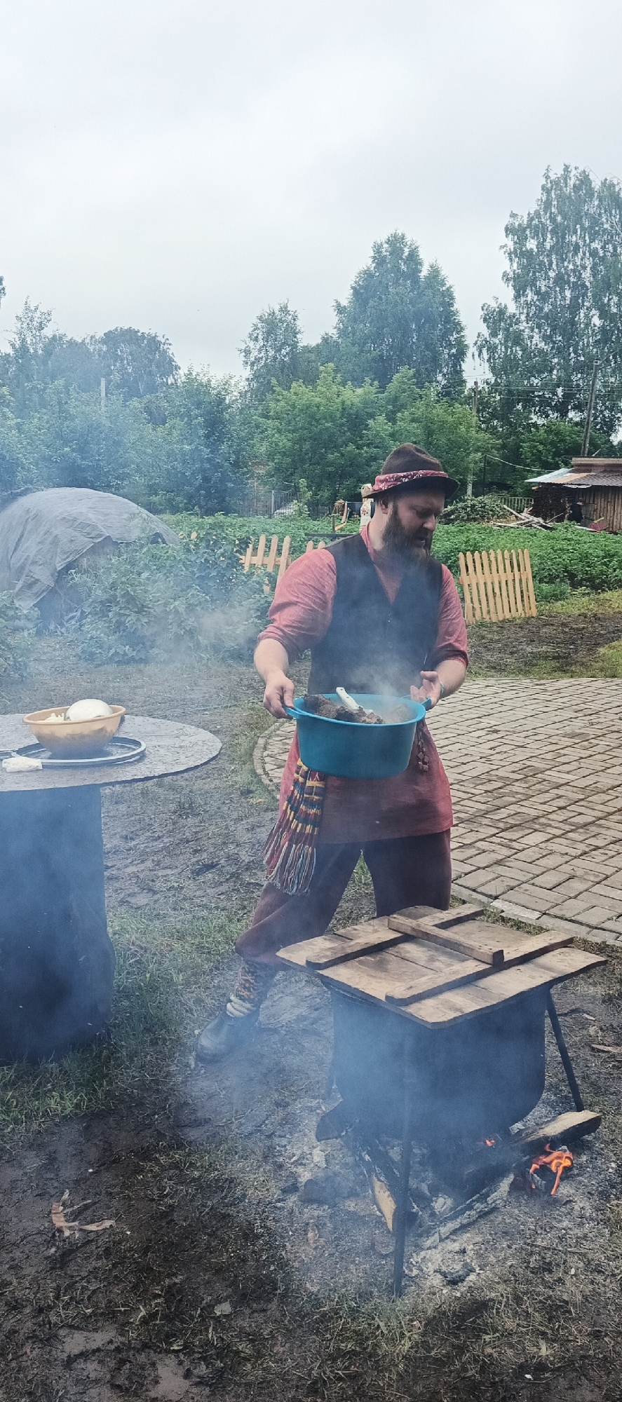 In the village of Starye Bygi, Russia, a man with a beard, wearing a red shirt, a dark vest, and a patterned headband, is preparing food outdoors. He holds a blue bowl containing pieces of meat. In front of him, a black cauldron rests on a stand over a fire, emitting a visible plume of smoke. To the left of the man, a round table holds a brown bowl filled with a light-colored substance, possibly food ingredients. The ground is a mix of grass and mud. In the background, trees, a wooden fence, and a covered structure are visible, suggesting a rural setting. The sky is overcast.