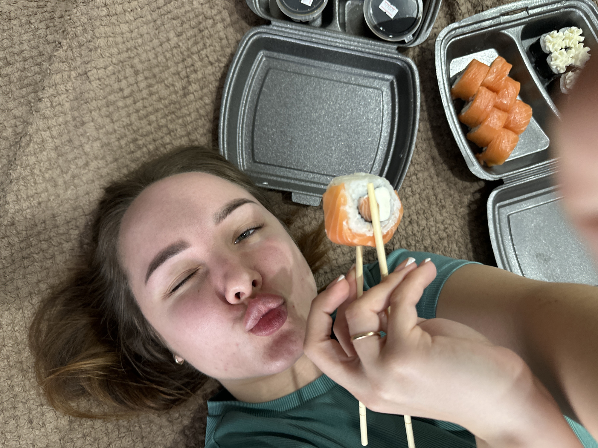 A woman is lying on a brown carpet, holding a piece of sushi with chopsticks. She is winking and puckering her lips. Next to her are two grey food containers and two small sauce containers. One container is empty and the other contains salmon rolls and some rolls with what appears to be cream cheese. The location cannot be determined based on the information in the image.