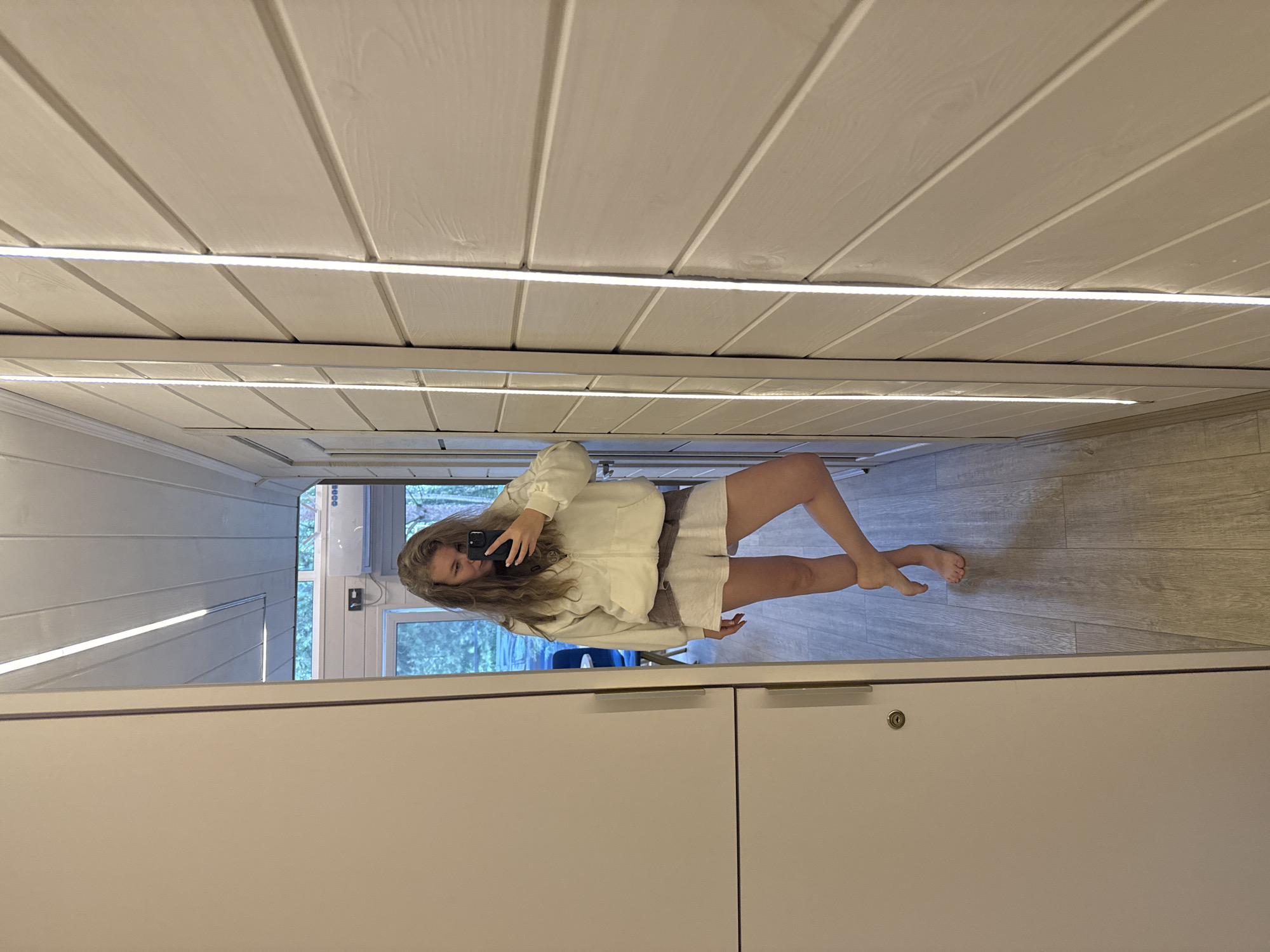 A woman is taking a selfie in a mirror. She is standing in a room with light wood-paneled walls and a wooden floor. The room appears to be in a modern home or apartment. The woman is wearing a white zip-up hoodie and light-colored shorts. Her hair is long and wavy. She is barefoot. The mirror is full-length and has LED strip lighting on either side. To the left of the mirror is a large white closet with a silver handle. The room appears to be clean and well-lit. The image was taken in Prokudino, Russia.