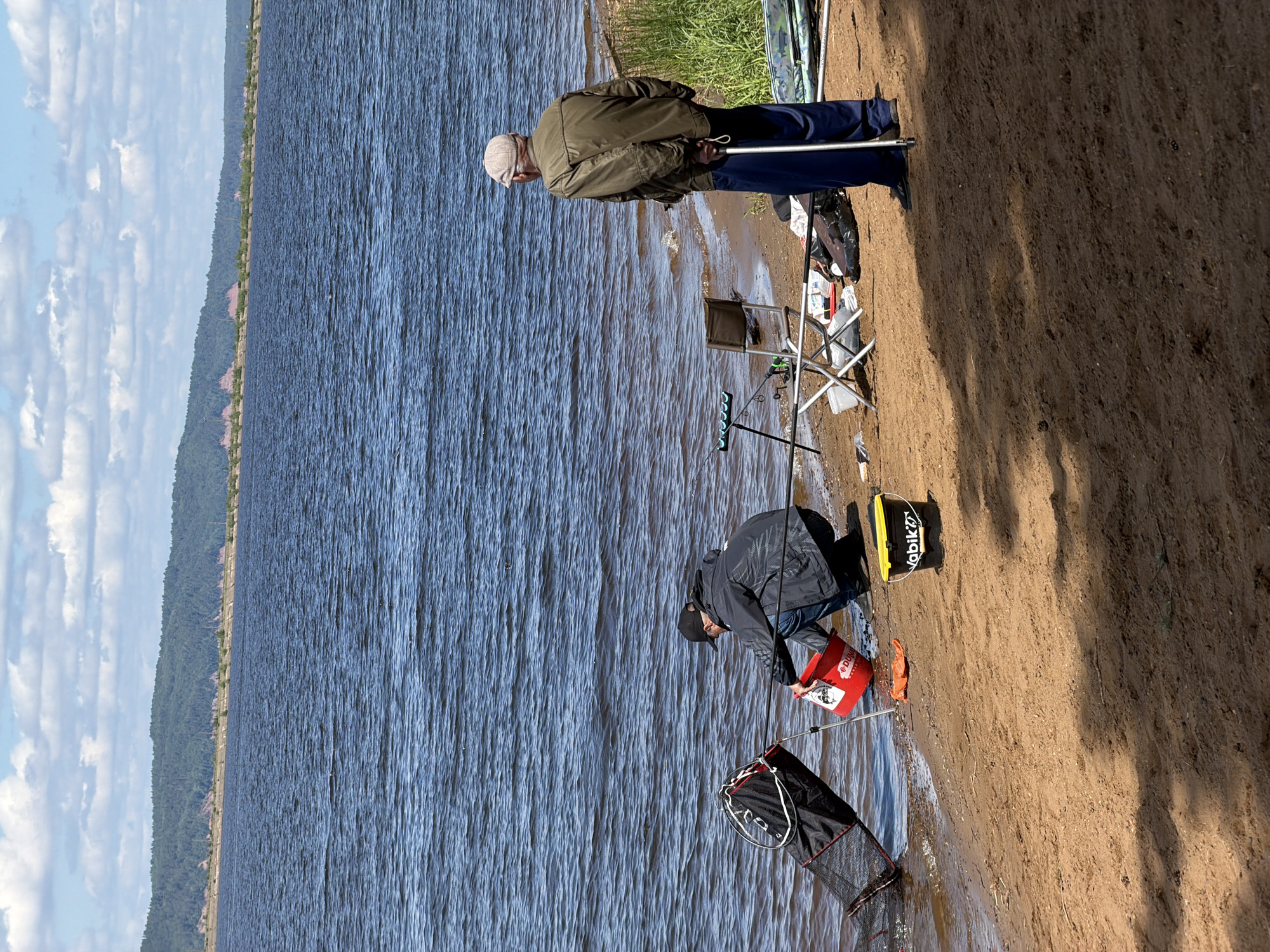 The image captures a serene outdoor scene, likely a fishing outing, on the bank of a large body of water, possibly a lake or wide river, with a distant forested coastline or hills under a partly cloudy sky. The setting is Chaikovsky, Russia.
Two men are present on the sandy, light brown shore. On the right, an older man, dressed in an olive-green jacket, blue trousers, and a light-colored cap, stands facing the water. He holds a long pole, presumably a fishing rod or support, and appears to be observing the water or his lines. Beside him, closer to the shore, is a silver folding chair, and various items of fishing gear are scattered on the ground, including a rod stand with multiple holders.
Further down the shore, closer to the water's edge, a younger man in a dark jacket, dark cap, and jeans is actively engaged in handling equipment. He is bent over a red bucket, which has the brand "DUNAEV" visible on its side. Partially submerged in the shallow water near him is a black mesh keepnet, likely for holding caught fish. Another yellow and black bucket, bearing the text "Vabik", sits on the sand nearby.
The water surface is rippled, reflecting the light from the bright daylight conditions. Shadows are cast on the sandy bank, suggesting it is midday or early afternoon. The overall atmosphere is calm and focused, indicative of a recreational fishing activity.