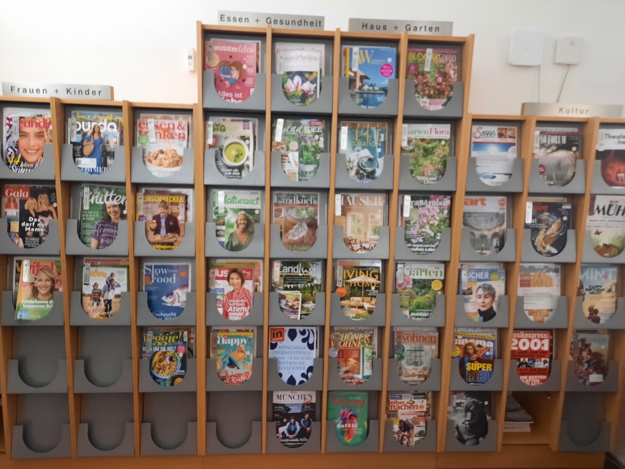 The image shows a magazine rack with several shelves filled with various magazines. The magazines are organized by category, indicated by signs above the shelves. The categories visible are "Frauen + Kinder" (Women + Children), "Essen + Gesundheit" (Food + Health), "Haus + Garten" (House + Garden), and "Kultur" (Culture). The magazine rack is made of wood and is located indoors, likely in a public space such as a library, community center, or store. The background wall is white. There are no people visible in the image. The magazines feature a variety of covers, with topics ranging from fashion and lifestyle to cooking, gardening, and entertainment. The overall impression is one of order and accessibility, with the magazines neatly displayed for browsing.