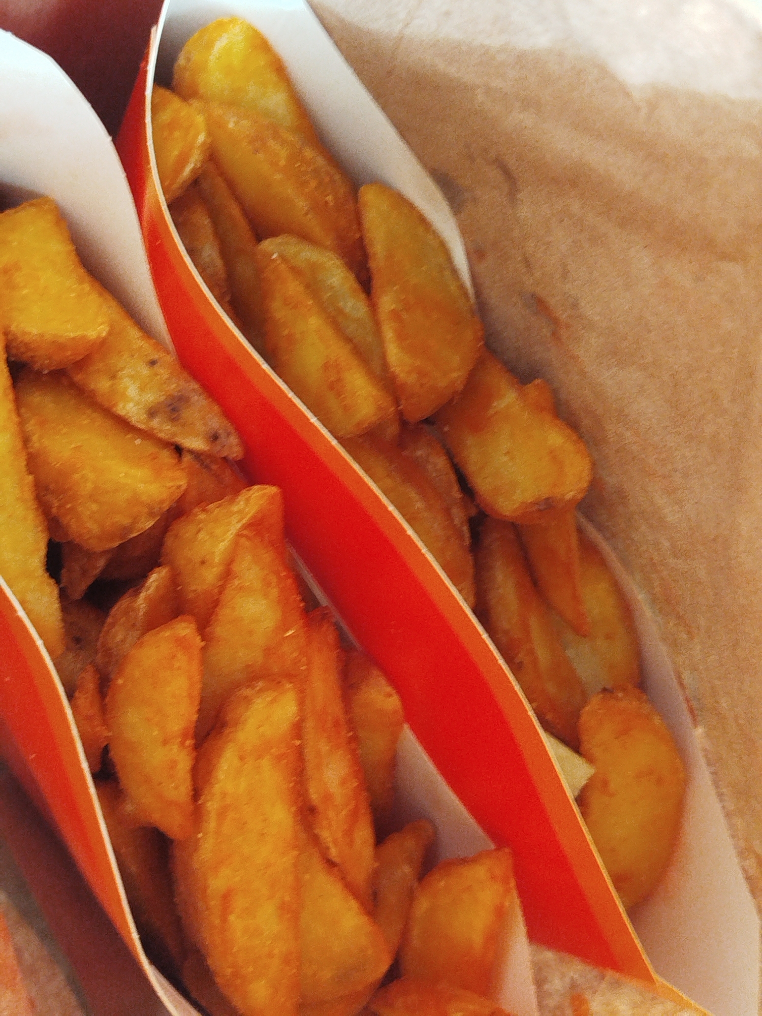 Two containers of potato wedges are visible inside a brown paper bag. The containers are made of white cardboard with an orange stripe. The wedges are golden brown, and some appear to have seasoning. The environment suggests a takeout or fast food setting. The image does not contain information to identify location.