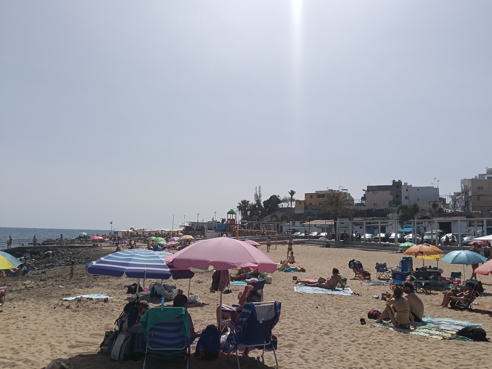 This is a photo of a beach in Mogán, Spain. The beach is sandy and there are many people sunbathing, swimming, and relaxing. In the foreground are several groups of beachgoers under umbrellas and in chairs. A couple sits on a towel. In the midground are more people on the beach, and a playground. The background features buildings, parking, and other infrastructure along the beachfront. The sky is overcast.