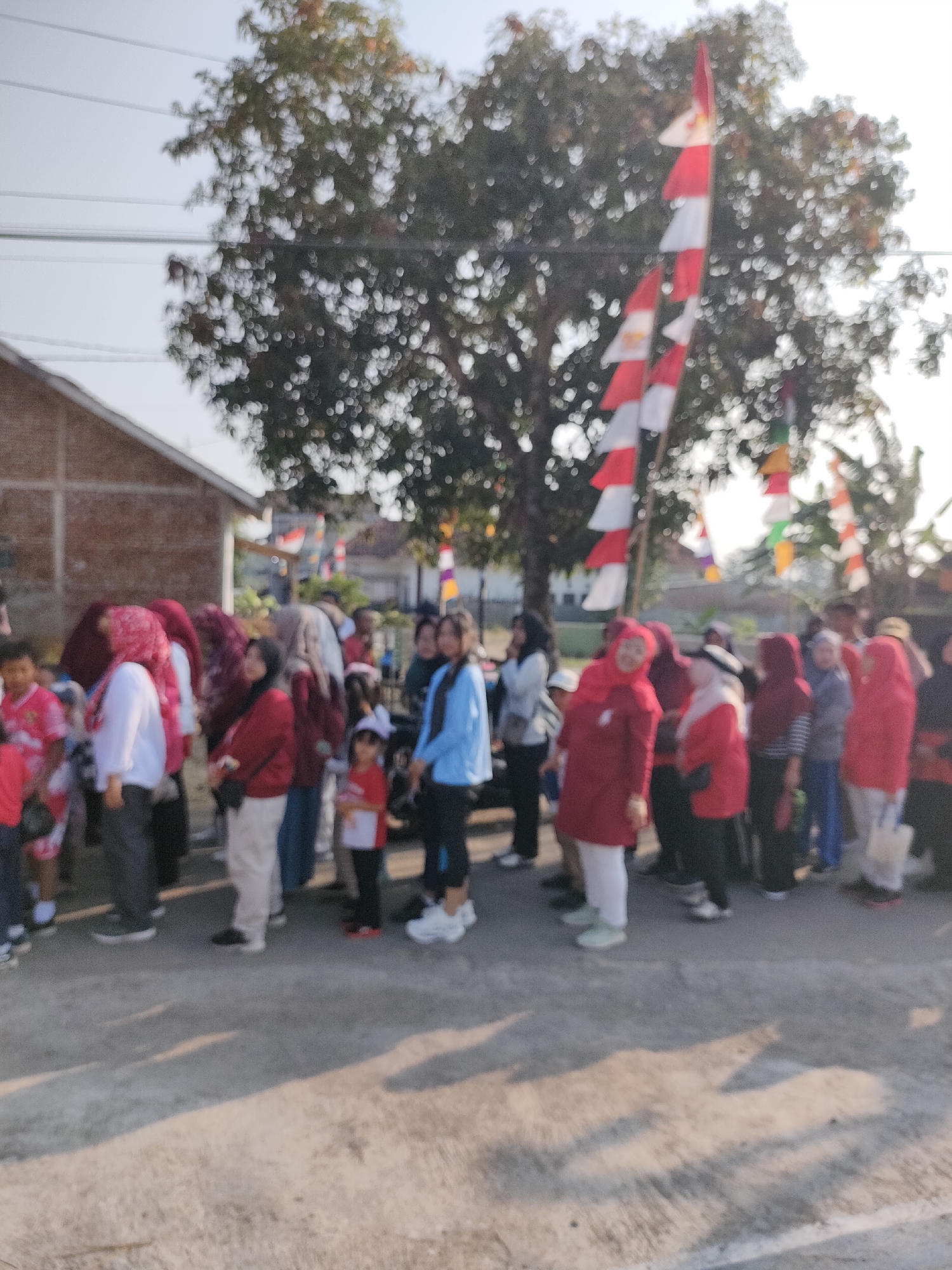 In this image, a group of people are gathered outdoors, possibly for an event or celebration. The people are of various ages and genders, and many are wearing red and white clothing, which could indicate participation in a patriotic event given the presence of Indonesian flags visible in the background. The setting appears to be a street or pathway lined with trees and a building. The lighting suggests it is daytime, possibly late afternoon due to the long shadows cast on the ground. The overall impression is one of community gathering, with a festive atmosphere implied by the flags.