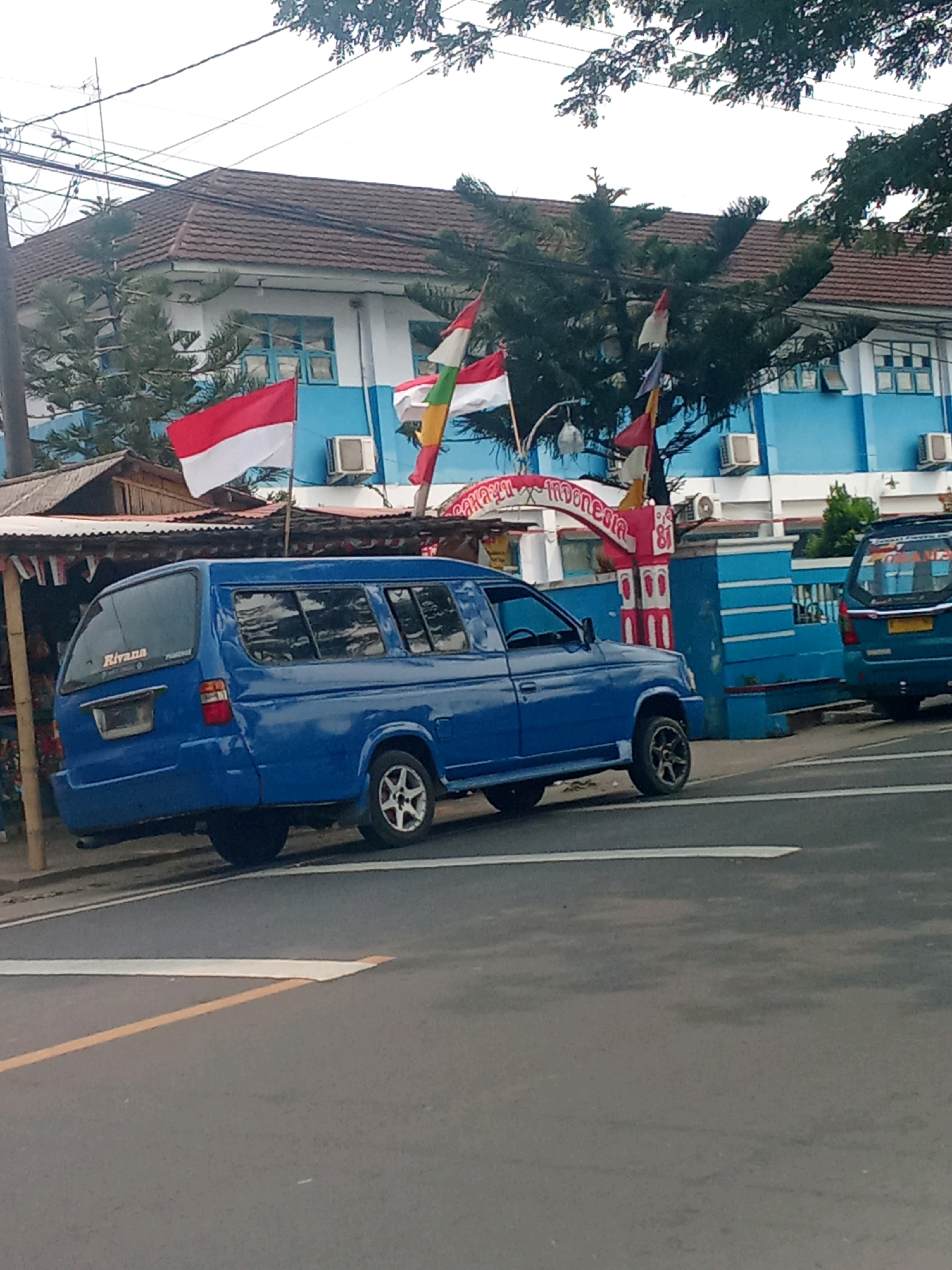 The image captures a street scene in Warunggunung, Indonesia, with a prominent blue van parked on the side of the road. In the background, a blue and white building with a brown tiled roof stands, adorned with Indonesian flags and other decorative elements. The building appears to be some kind of institution, possibly a school or government office, indicated by air conditioning units visible on its exterior. A white and red archway with text, likely in Indonesian, is situated in front of the building. Another blue vehicle, possibly a different model of van, is parked to the right of the main subject. The foreground shows a paved road with white and yellow lane markings. The scene is framed by trees, suggesting an outdoor, public space. The lighting indicates it is daytime.