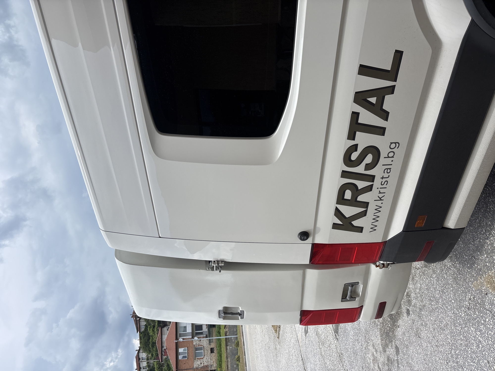 The media file shows the rear of a white van with the text "KRISTAL" and "www.kristal.bg" printed vertically in dark grey. The van has two rear doors, with a latch visible on the left door and a handle on the right door. Red taillights are present on both sides of the rear bumper. The van is parked on an asphalt surface, likely a street or parking lot, with a grey, cloudy sky overhead. In the background, buildings with red tile roofs and greenery are visible, suggesting a town or village setting. The lighting and cloudy sky indicate it is daytime, possibly late afternoon or early morning. The overall scene is static, with no people or discernible activity. The location is identified as Hvoina, Bulgaria.