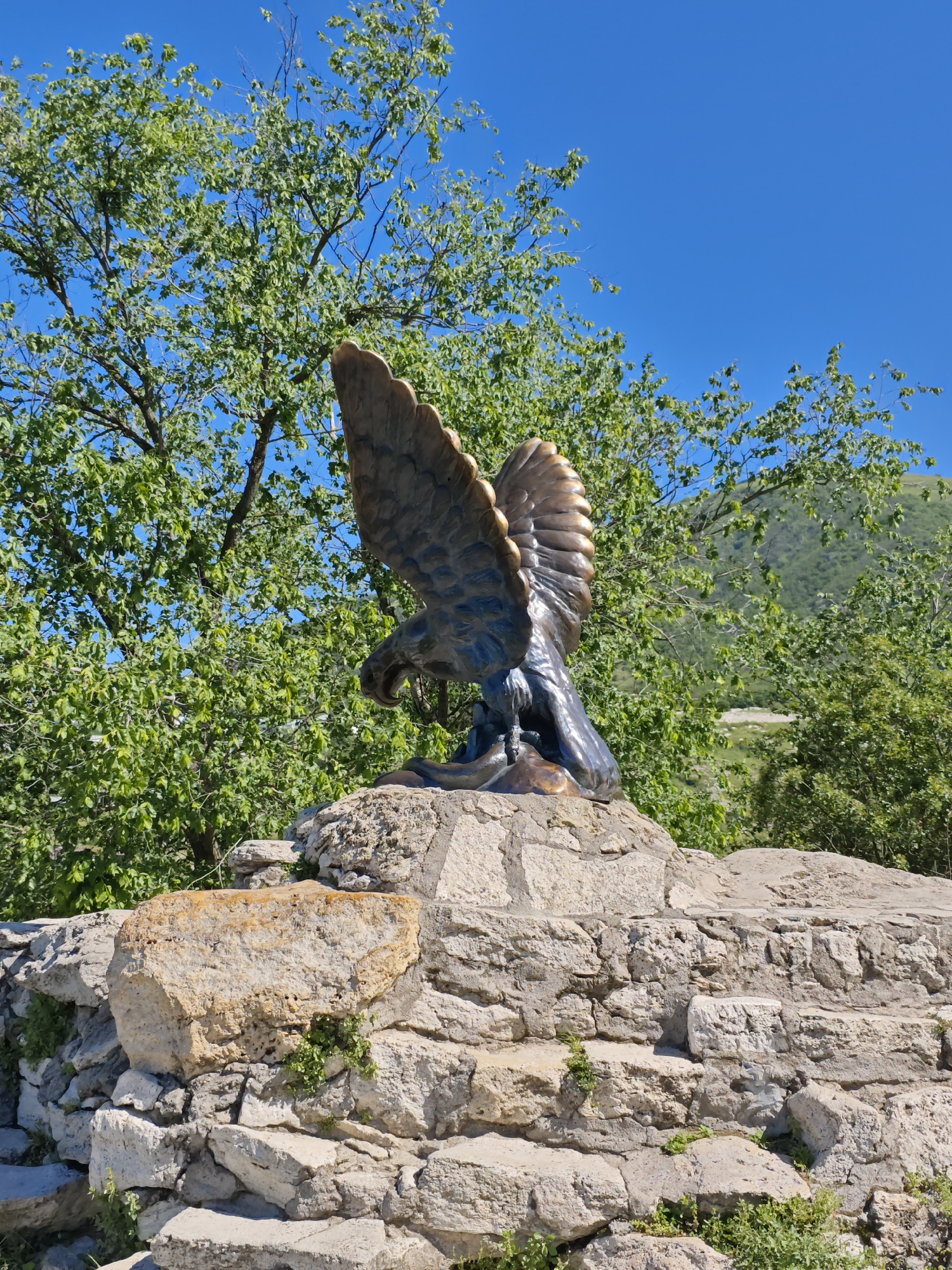 A bronze statue of an eagle stands prominently on a rustic stone structure under a clear blue sky. The eagle, positioned with its wings partially spread, appears to be either landing or preparing for flight, and its head is turned downwards with a slightly open beak. The statue exhibits a dark, metallic finish, possibly bronze, with lighter, patinated areas highlighting its textured feathers.
The eagle is perched on a robust foundation made of irregularly shaped, light-colored stones, some displaying warm, yellowish hues. Small green plants and foliage can be seen growing between the cracks of these stacked rocks, enhancing the natural feel of the monument's base.
In the background, lush green trees with dense foliage and visible branches create a vibrant backdrop. Beyond these trees, gentle green hills or mountains rise under a vast, cloudless blue sky. The scene is bathed in bright sunlight, indicating it is daytime, likely morning or early afternoon, with clear and sunny weather conditions. There is no visible text in the image. The setting evokes a scenic, elevated outdoor location, characteristic of the landscapes found in Pyatigorsk, Russia.