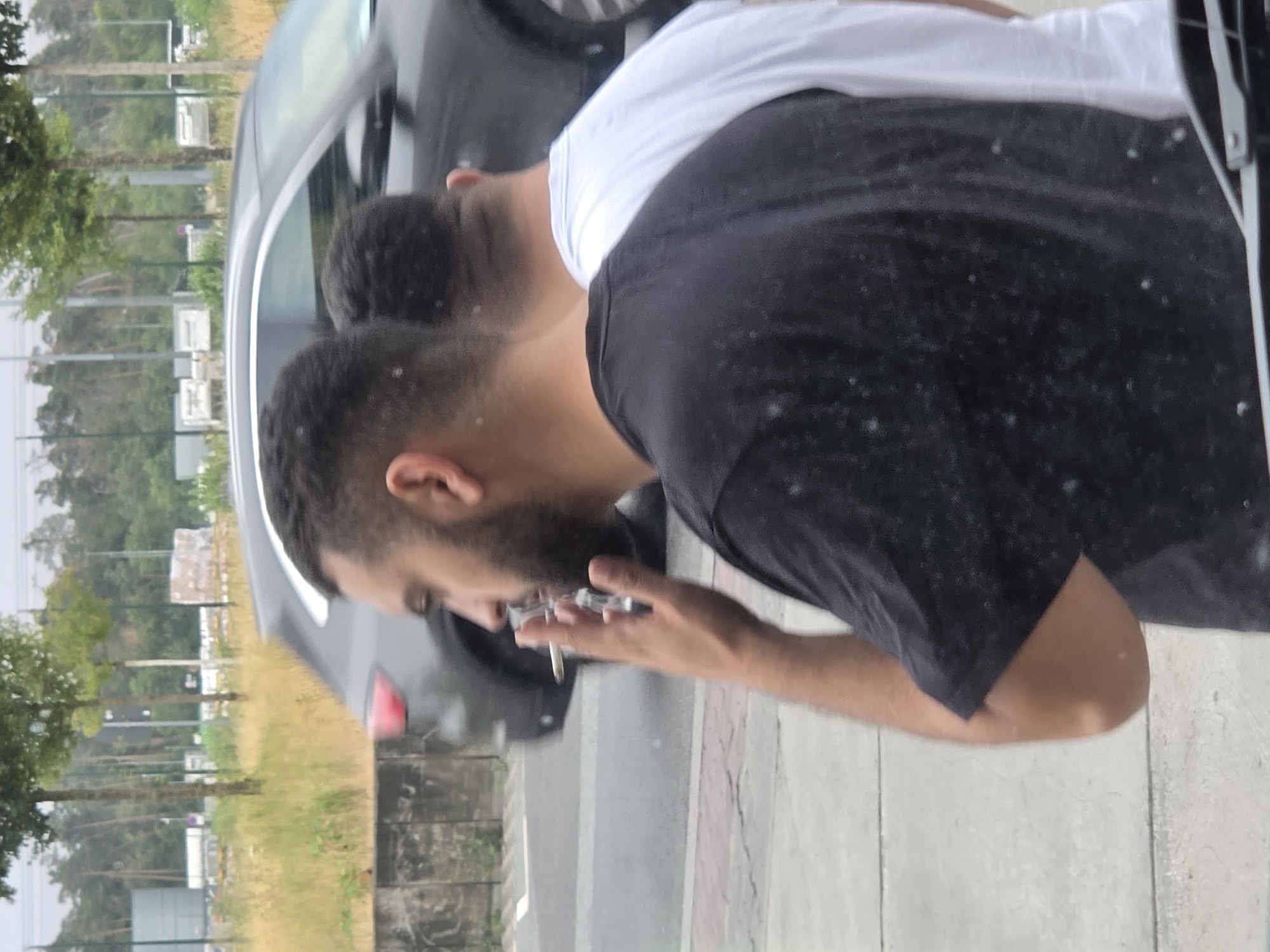 The image shows a side profile of two men standing on a sidewalk. The man in the foreground, on the left, is wearing a black t-shirt and has short dark hair and a beard. He is holding a lit cigarette to his mouth. The second man, partially visible behind him and to the right, is wearing a white t-shirt and has short dark hair. They are standing in front of a dark-colored car. The background includes a sidewalk, a road with a car driving on it, a low concrete wall, and a grassy area with trees and some industrial-looking structures in the distance. The scene appears to be outdoors, possibly in an urban or semi-urban environment. The image quality is moderate, with some grainy texture and a slightly overcast sky. The location is identified as Walldorf, Germany.