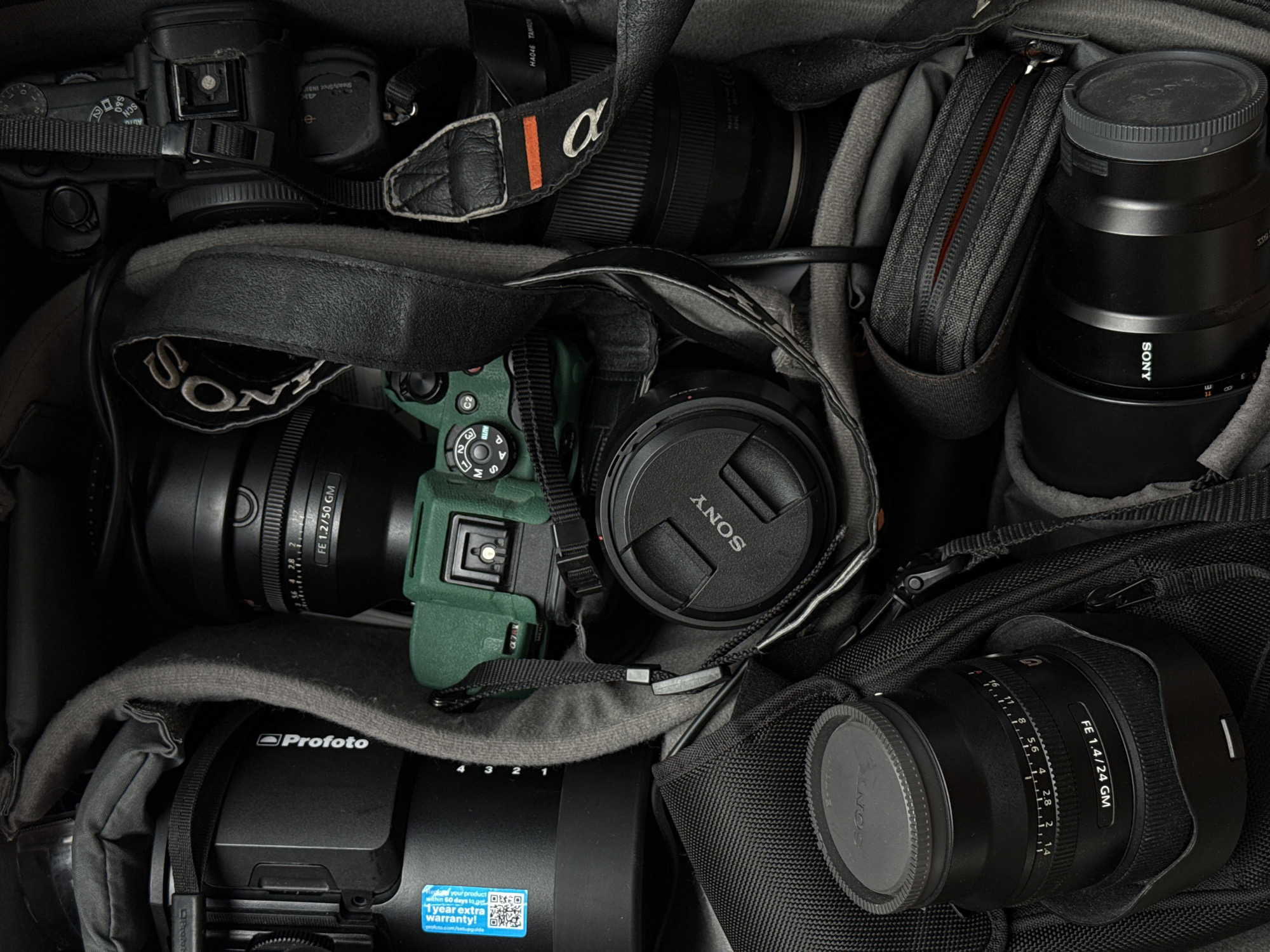 The image displays a top-down view of various professional camera equipment neatly packed within a grey camera bag or insert, suggesting either storage, transport, or preparation for a photography session. The predominant colors are black and grey, with one camera body featuring a distinct green protective casing.
Key subjects include multiple Sony mirrorless camera bodies, several high-end lenses, and a Profoto lighting accessory. Visible lenses include a Sony FE 1.2/50 GM and a Sony FE 1.4/24 GM, indicating a preference for fast prime lenses, along with a lens partially identified by "HA046 TAMRON."
Notable details include:
* One camera body's mode dial displays settings such as "SCN," "S&Q," and "Auto." Another camera body shows "4K" and "SteadyShot INSIDE."
* The green-cased camera features a mode dial with "AUTO," "M," "S," "A," "P," "C1," and "C2" settings.
* "SONY" logos are visible on multiple lens caps and camera straps, alongside an "alpha" logo.
* A larger black unit is identifiable as "Profoto," bearing a blue sticker that reads "Register your product within 60 days to get 1 year extra warranty!" with a QR code and the URL "profoto.com/webguide."
* The equipment appears well-maintained and organized within padded dividers.
No specific geographical or environmental cues are discernible from the image itself. The equipment suggests professional-grade photography gear.