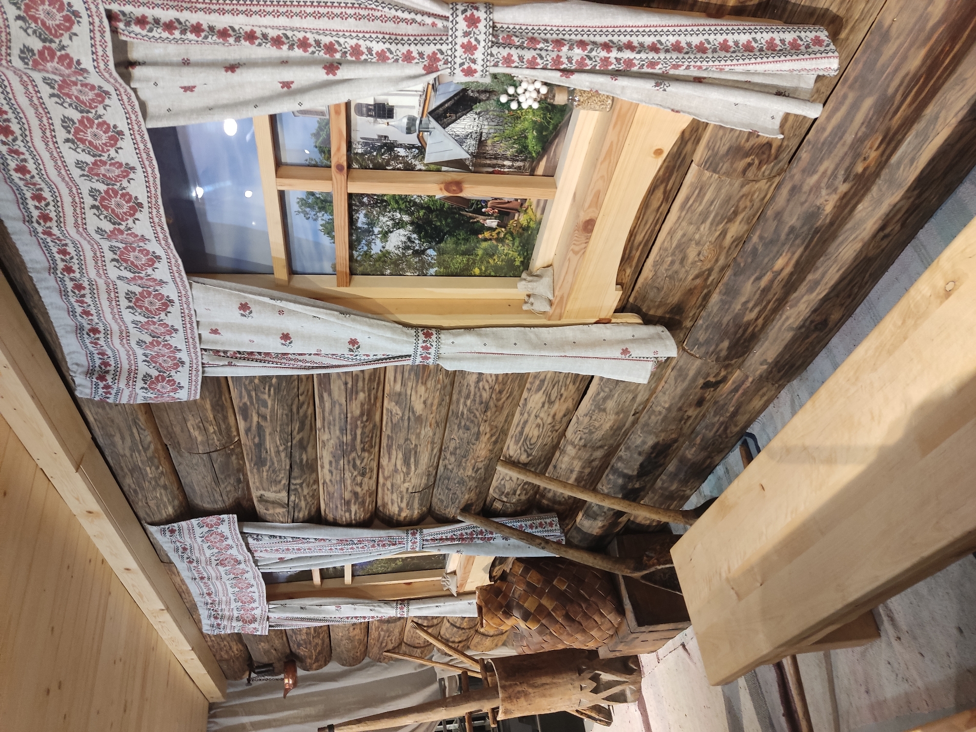 The image shows the interior of what appears to be a rustic building, possibly located in the city of Pskov, Russia. The ceiling is constructed of rough-hewn wooden logs. Two windows are visible, adorned with curtains that feature a traditional floral pattern in red and gray. The view through the windows reveals a garden or yard scene, with trees and buildings in the background. A wooden basket and other rustic objects are present, adding to the traditional aesthetic. The walls of the room are made of light-colored wood planks.