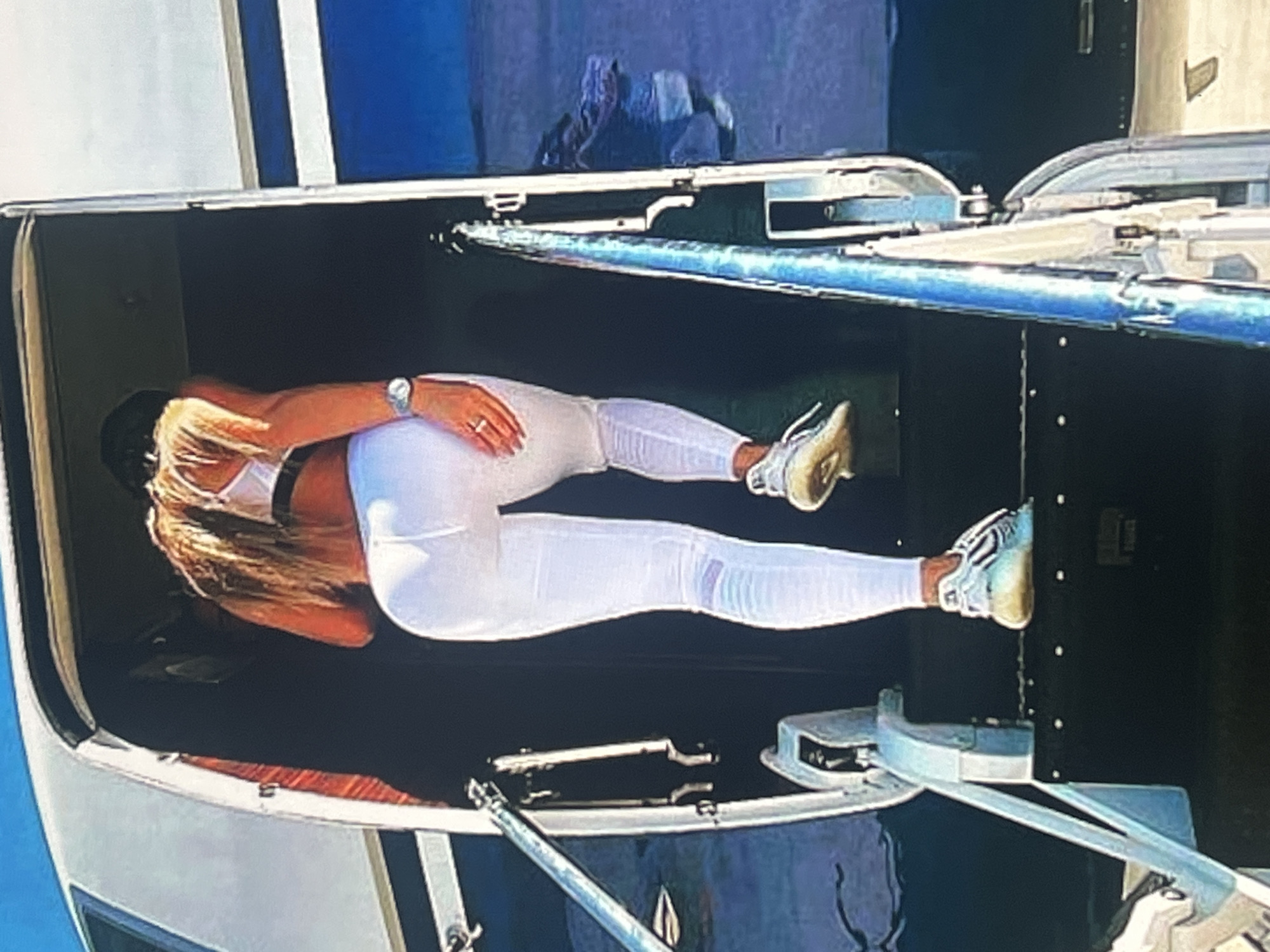 A person is walking up the stairs of an airplane. They are wearing white leggings, a white sports bra, and white sneakers. The airplane is white with blue accents. The background is a clear blue sky. The scene appears to be on an airport tarmac. There is no information in the image to suggest it is in Vanadzor, Armenia.