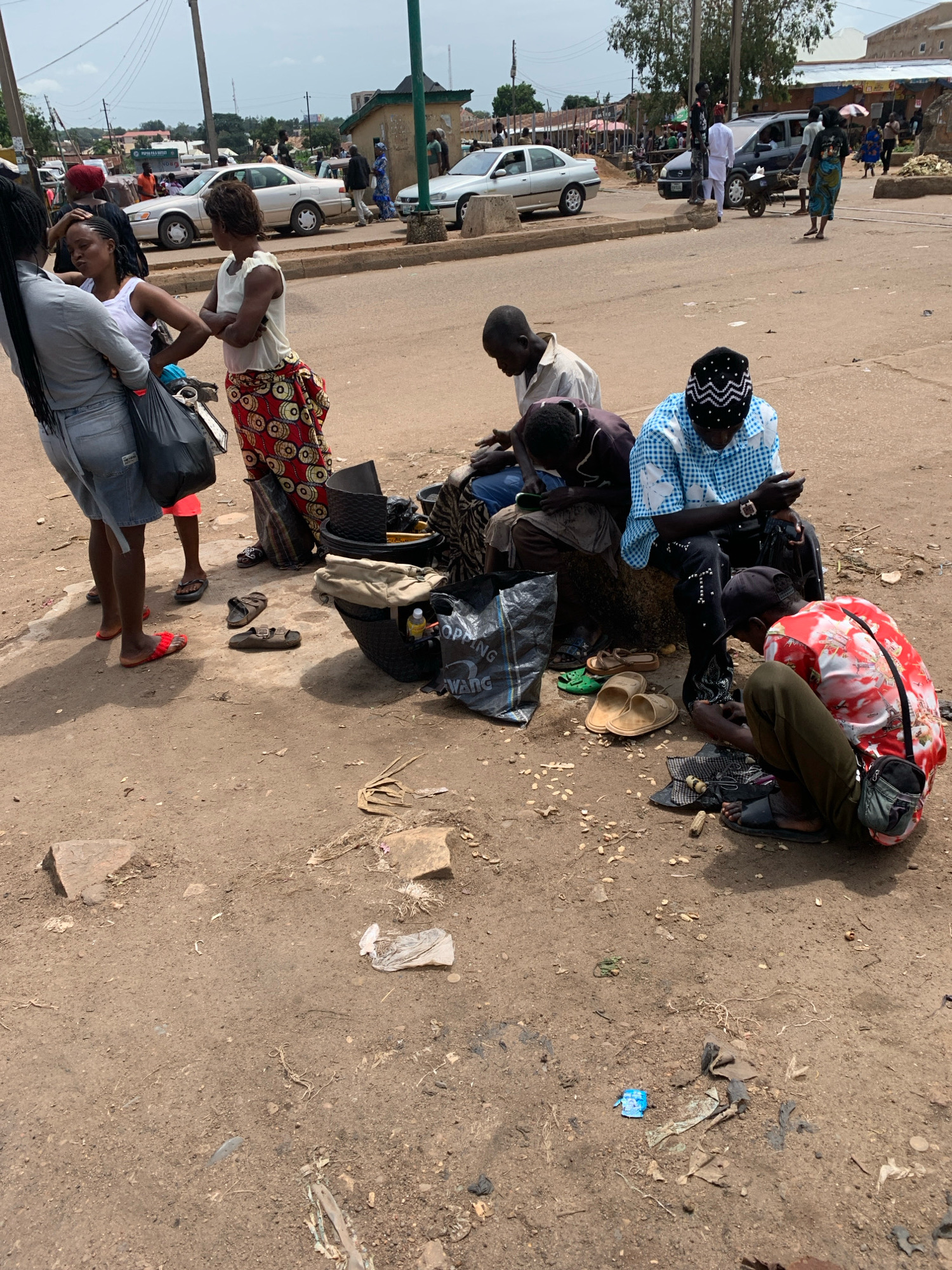 In Bukuru, Nigeria, a dusty, sun-drenched street scene unfolds. Several individuals are gathered, some engaged with mobile phones while others appear to be resting or waiting. The ground is unpaved and littered with debris, including plastic bags, discarded food items, and various small objects. In the background, a few vehicles, including cars and a motorcycle, are visible on the road, along with market stalls and buildings that line the street. The overall atmosphere suggests a busy, everyday street in a Nigerian city.
