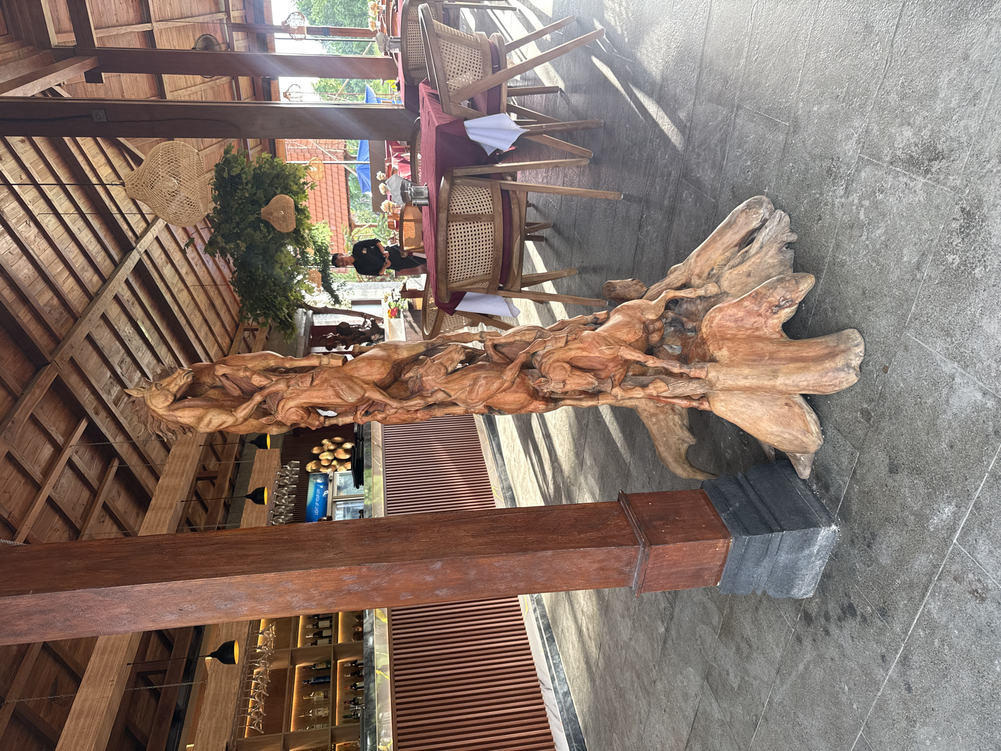 This image shows the interior of a restaurant in Ubud District, Indonesia. In the foreground, a large, intricately carved wooden sculpture of horses stands prominently. The sculpture is made from a single piece of wood, with the horses appearing to be in motion, some rearing and others running. The wood has a natural, rustic finish, highlighting the grain and texture. The sculpture is supported by a base of gnarled roots.
Behind the sculpture, a wooden bar area is visible, with shelves stocked with bottles and glassware. The bar is illuminated by pendant lights. Further in the background, dining tables are set with red tablecloths and white napkins. Cane-backed chairs surround the tables. A person in a black uniform is standing near the tables, possibly a waiter or staff member.
The restaurant features a thatched roof supported by wooden beams, characteristic of traditional Balinese architecture. Large windows or open spaces allow natural light to enter, creating a bright and airy atmosphere. Hanging woven lamps add to the ambiance. Lush green foliage is visible in the background, suggesting an outdoor or garden setting beyond the restaurant. The floor is paved with gray stone tiles. The overall impression is one of a warm, inviting, and culturally rich dining establishment.
