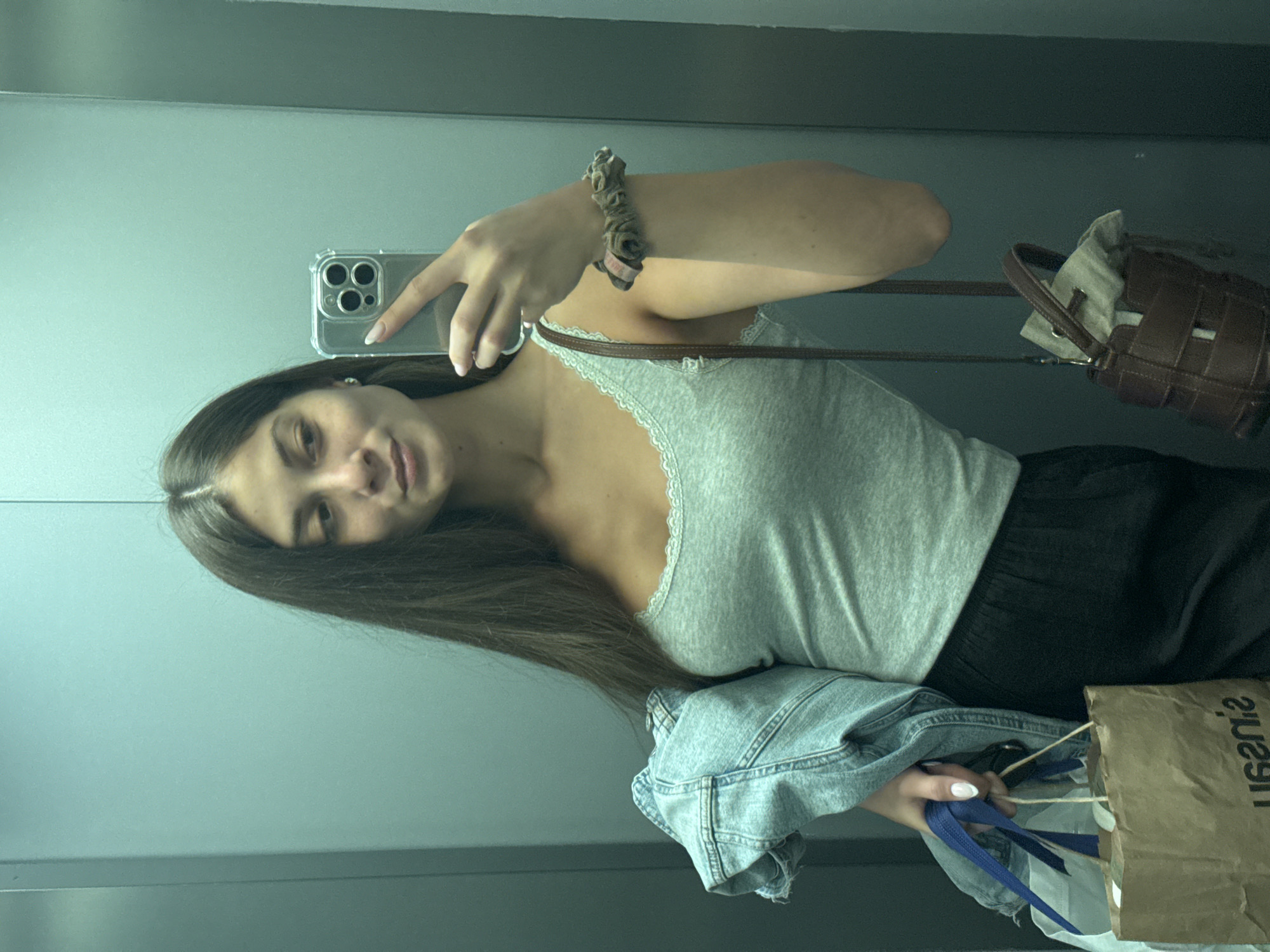 The image is a mirror selfie, rotated 90 degrees clockwise, depicting a young woman in an indoor setting with plain, light-colored walls. The woman has long, straight dark brown hair, parted in the middle, and is looking towards the camera with a neutral to slight smile.
She is wearing a light grey tank top with white lace trim and black pants or a skirt. A light blue denim jacket is casually draped over her arm. On her right wrist, she wears a brown scrunchie. A small, dark brown woven-style handbag with a drawstring closure hangs over her left shoulder. In her left hand, she holds a smartphone (an iPhone with a clear case and a triple camera array) to take the selfie, revealing a French manicure on her nails. Her right hand is holding a brown paper shopping bag from which blue ribbons or fabric pieces are visible. The paper bag features reversed text, which appears to spell "ISANA" or "LISANA" when read forwards.
The background consists of plain, light-colored walls, possibly grey or light blue, suggesting an indoor space like an elevator or hallway. The lighting is uniform, indicating artificial light, so the time of day is indeterminate. The location context provided is Burgas, Bulgaria.