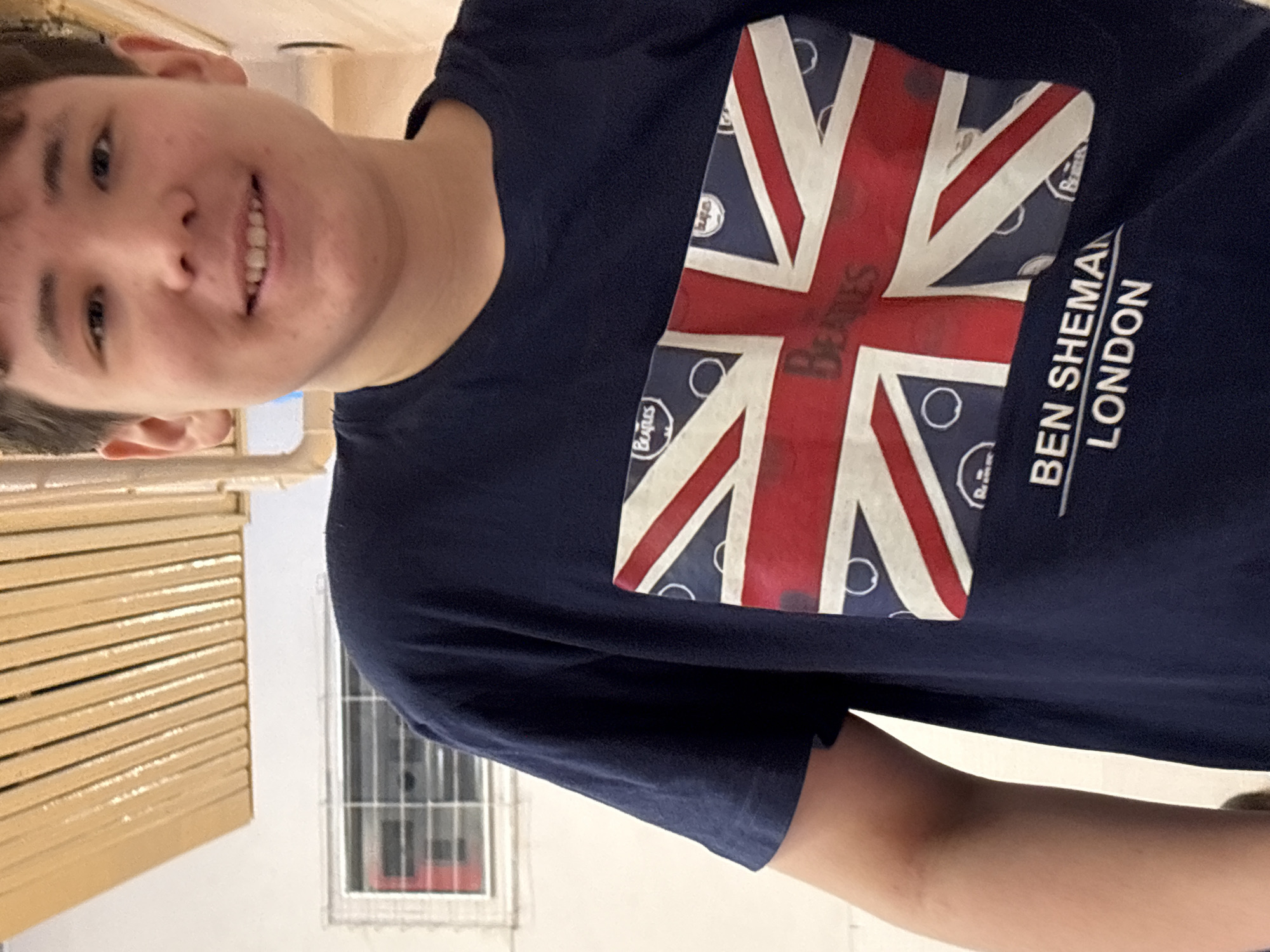 A young person is pictured wearing a navy blue T-shirt with a British flag design that incorporates the name "Beatles" and text reading "BEN SHERMAN LONDON." The person is smiling and appears to be indoors. The background includes a window and a set of wooden drawers. The location is not discernible from the image.