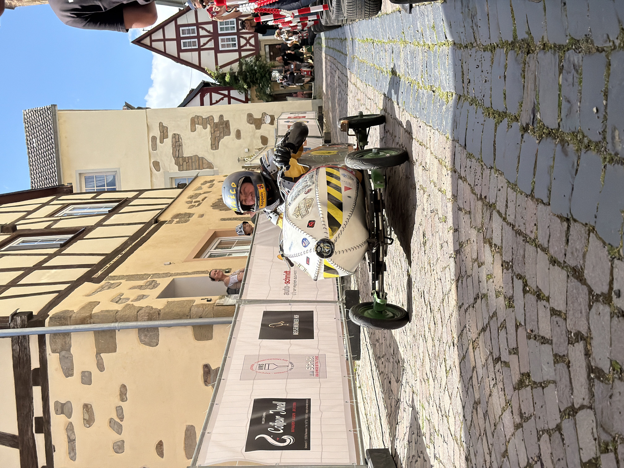 The image depicts a gravity-powered vehicle, likely a soapbox racer, descending a cobblestone street in Meisenheim, Germany.
In the foreground, a person wearing a black full-face helmet with "NICO" and "KAPPELN" visible, protective yellow and grey/metallic attire, and gloves, is seated within a custom-built vehicle. The vehicle features a white, spheroid front section with black stitching-like outlines and yellow and black hazard stripes, along with various small stickers. The visible front wheels have green hubs and black tires. The driver maintains a slight smile while gripping an internal steering mechanism.
The road surface is composed of irregular cobblestones, with sparse green vegetation growing between the stones. Sunlight illuminates the left side of the street, while the right side is partially in shadow.
To the immediate left of the vehicle, temporary white barriers are erected, displaying four sponsor advertisements: "Coiffeur Janell," "WMS RAUMGESTALTUNG," "MEISENHEIMER HOF" (confirming the city location), and "auto-schmidt." These barriers are anchored by grey concrete blocks. Behind the barriers, an individual in a hat is partially visible. Further back, a woman in a light-colored top leans from an open window or balcony of a building, observing the scene with a smile. The buildings on the left are multi-story, featuring light-colored stucco facades, exposed timber framing, and some decorative stonework. A metal downspout runs vertically on one building.
On the right side of the street, a makeshift barrier of stacked black tires and red-and-white striped plastic bollards delineates the track. Further down the street, multiple spectators are visible behind these barriers. The background features traditional German half-timbered houses with white walls, dark timber frames, and tiled roofs, set against a clear blue sky with minimal white clouds.
The overall scene suggests a community event or race taking place on a sunny day.