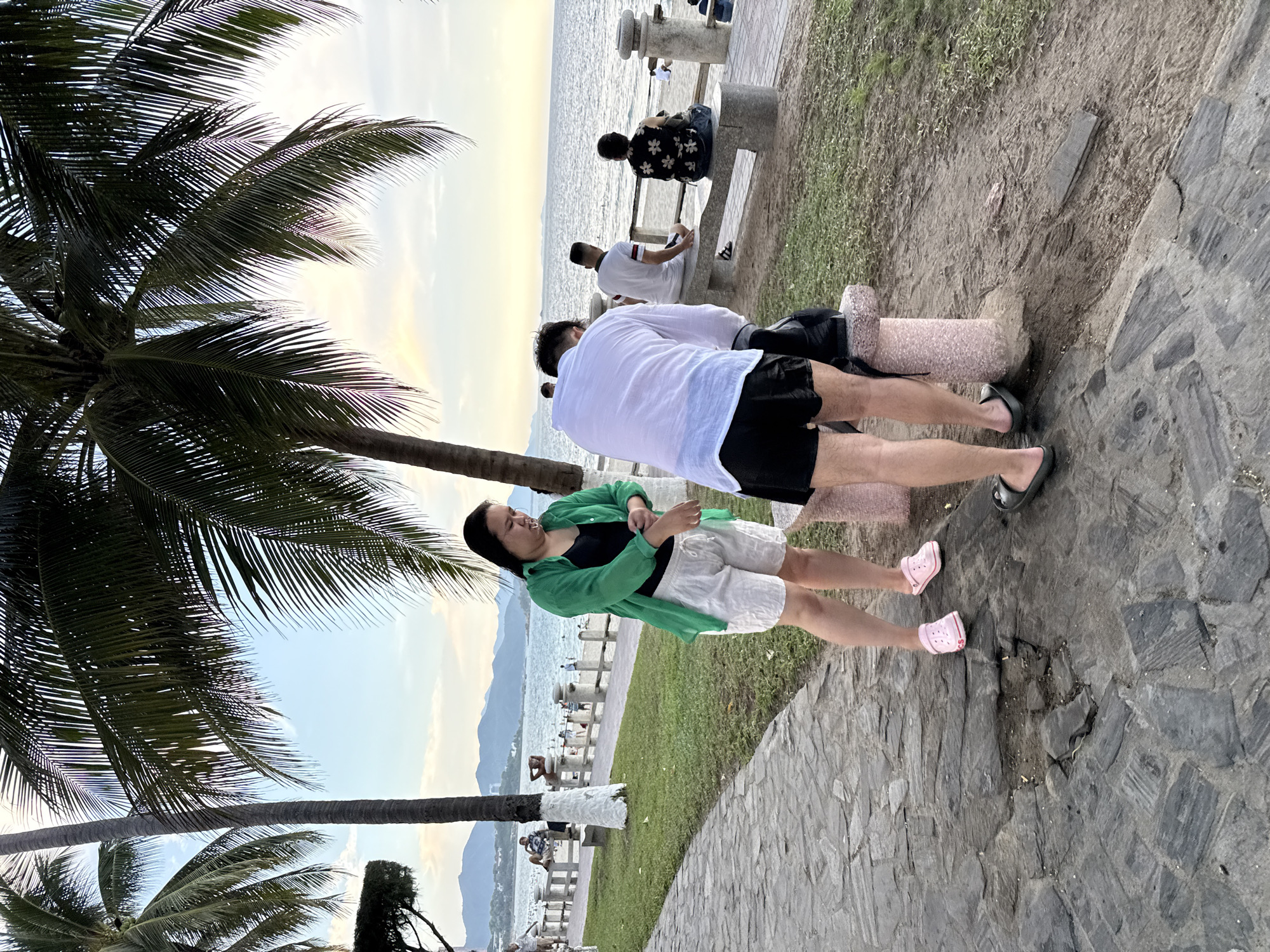 The image depicts a coastal pathway in Nha Trang, Vietnam, likely along Tran Phu Boulevard, characterized by a stone walkway and grassy areas bordering a beach. Two individuals are in the foreground: a woman in a green open-front shirt, black tank top, and white shorts, with pink Crocs, is adjusting the sleeve of her shirt. Next to her, a person in a white button-down shirt, black shorts, and dark flip-flops, leans over a stone bench, appearing to adjust something on their leg or backpack. In the background, other people are seated on benches facing the ocean, and the sea meets a sandy beach under a hazy sky with mountains in the distance. Palm trees frame the scene, their fronds casting shadows.