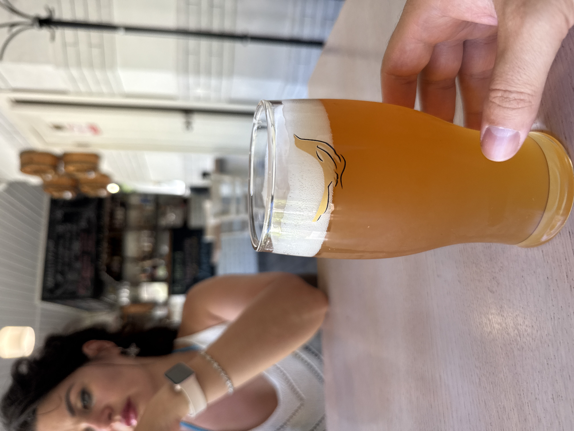 A close-up shot captures a glass of amber-colored beer with a frothy head, sitting on a light-colored wooden table. A hand, with fingers curled around the base of the glass, is visible on the right side of the frame. The beer glass has a small, black outline of a bird on it. In the background, out of focus, a woman with dark hair is seated at another table, wearing a white top and a watch on her left wrist. Behind her, shelves with bottles and other items are visible, suggesting a bar or restaurant setting. The overall lighting indicates it is daytime. The environment appears to be a casual dining establishment. There are no explicit visual cues to pinpoint the location to Sochi, Russia.
